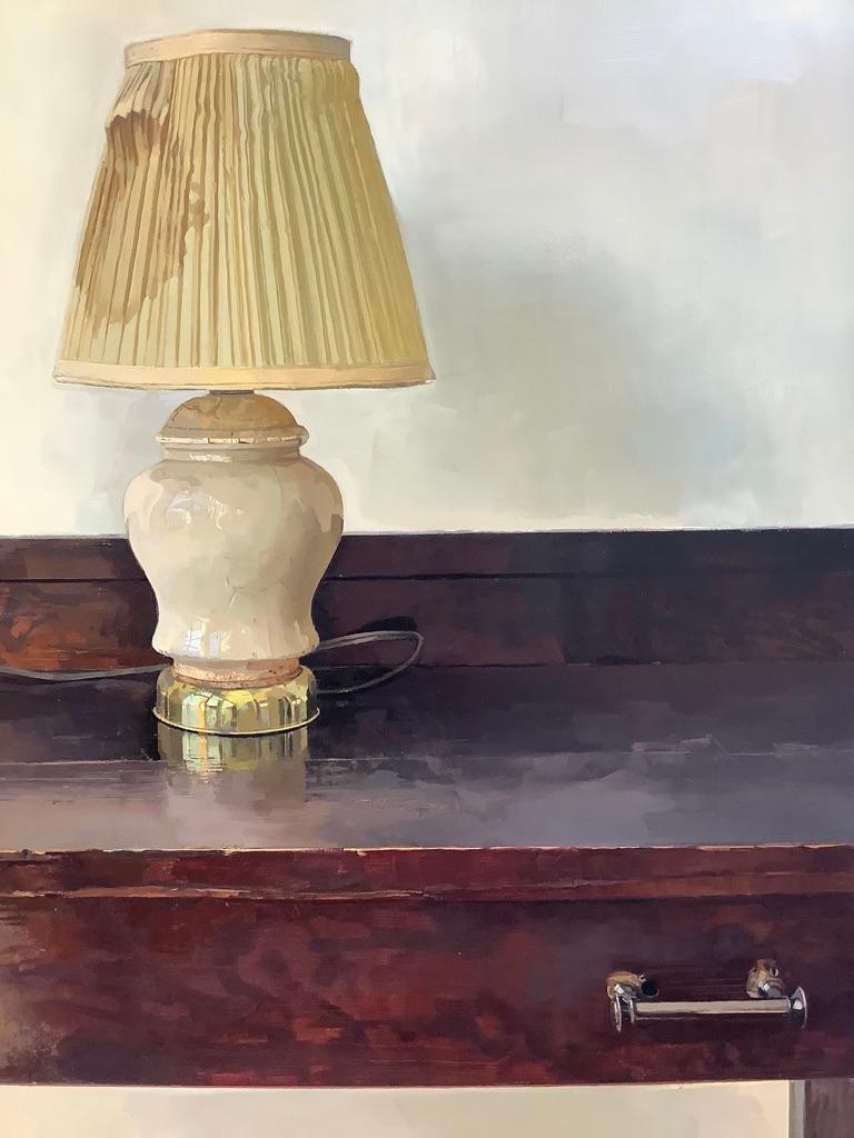 In diesem ruhigen Stillleben steht eine einzelne Lampe mit einem vergilbten Lampenschirm auf einem dunkelbraunen Mahagoni-Holzschreibtisch mit grauen Metallgriffen an den Schubladen. Das Objekt und die Möbel wirken unerwartet dramatisch vor einer