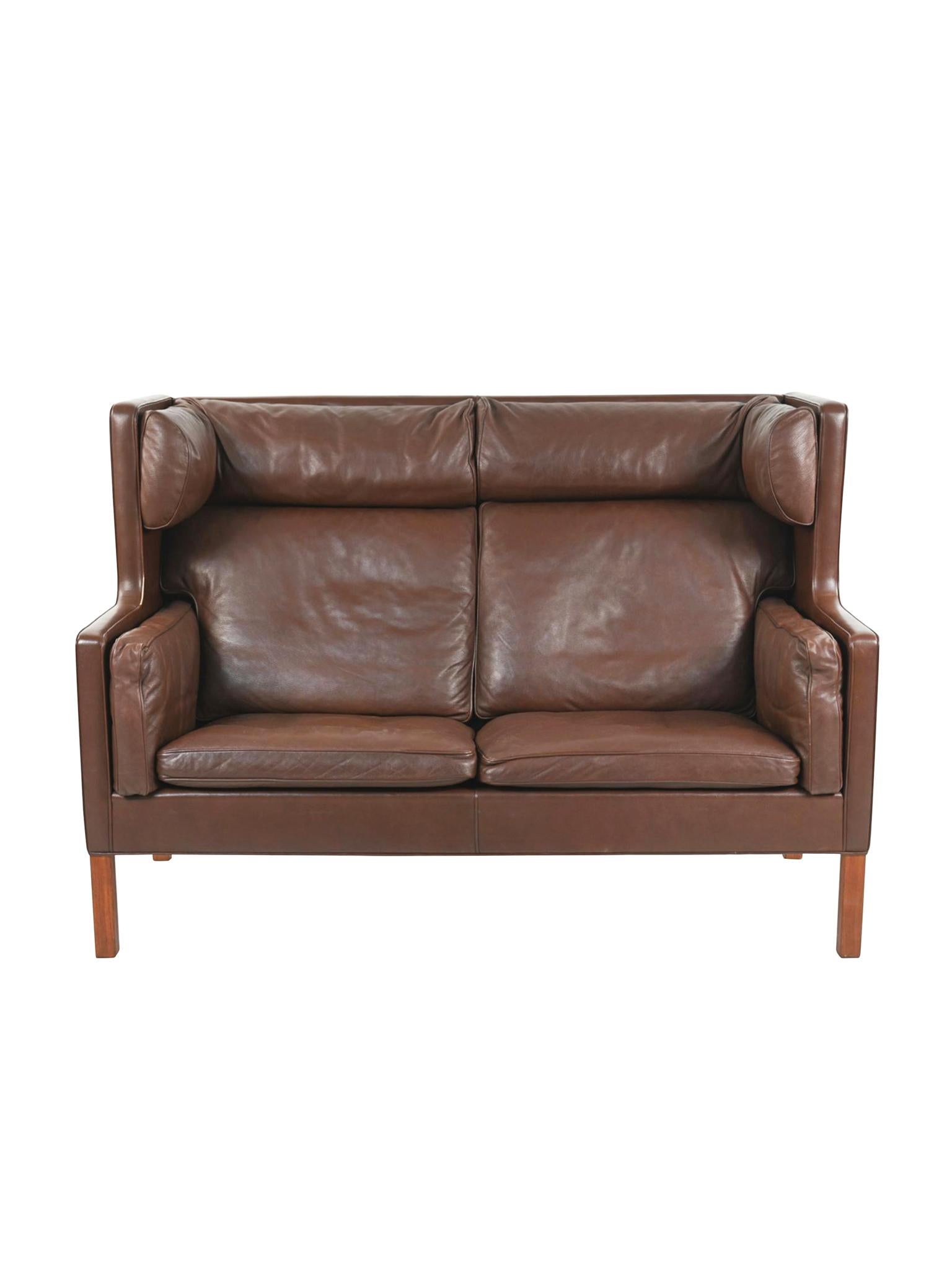 This semi-aniline brown leather coupé sofa was designed by Børge Mogensen for Fredericia in the 1960s. Structurally, the sofa has a high, cushioned back, reminiscent of a wingback armchair, inviting an upright but relaxed position. While the front