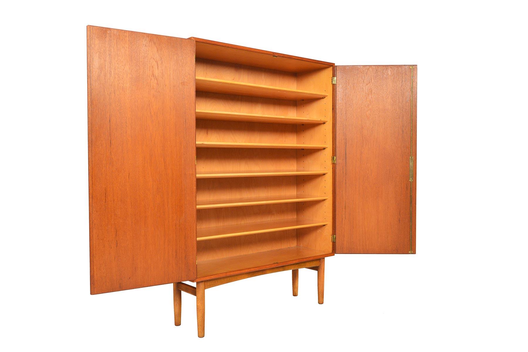 This narrow, tall cupboard was designed by Børge Mogensen for Søborg Møbelfabrik in the 1950s. Two large teak cabinet doors with the designer's signature pulls open to reveal a spacious birch lined interior with six adjustable shelves. Case stands