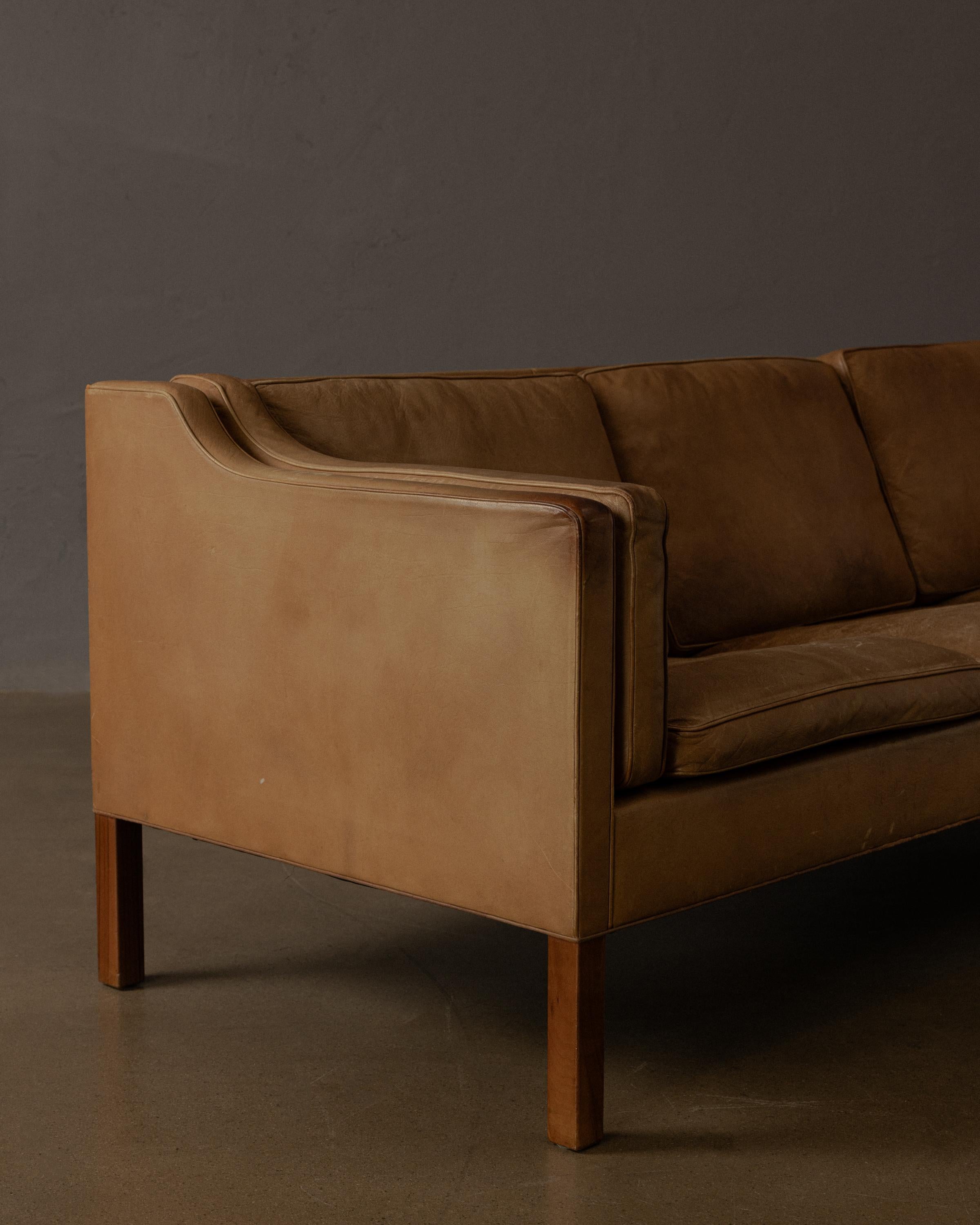 A three-seat sofa designed by Børge Mogensen, crafted in Denmark in the 1950s. This piece features clean, architectural lines and subtle curves at the armrests, upholstered in richly patinated, natural leather that has aged over time, it rests on