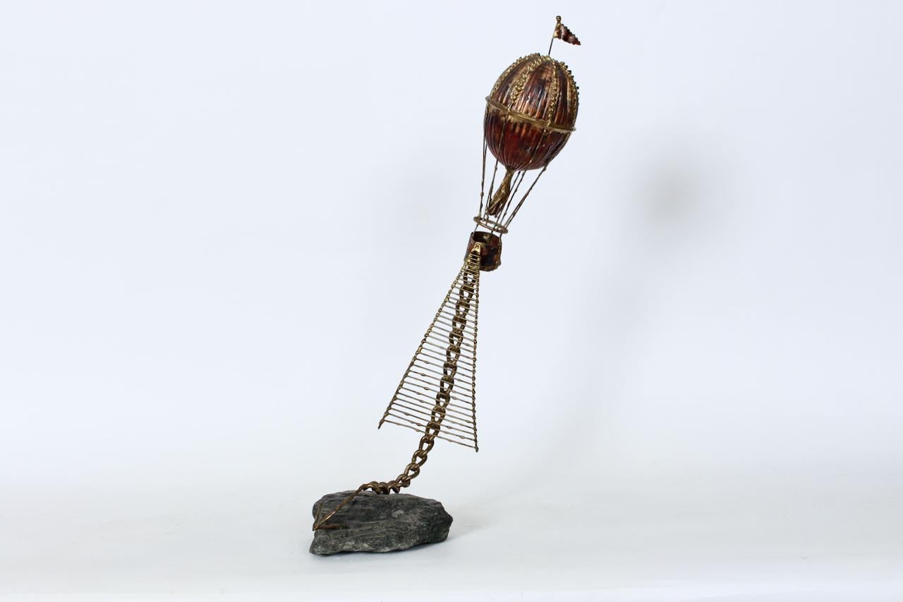 Scultura da tavolo a tecnica mista con mongolfiera di Brian Bijan. Una scultura artigianale in metallo misto saldato in rame, ottone rivestito di ferro e basalto, con pallone e cesto in rame stile inizio '900, bandiera in ferro saldata in ottone,