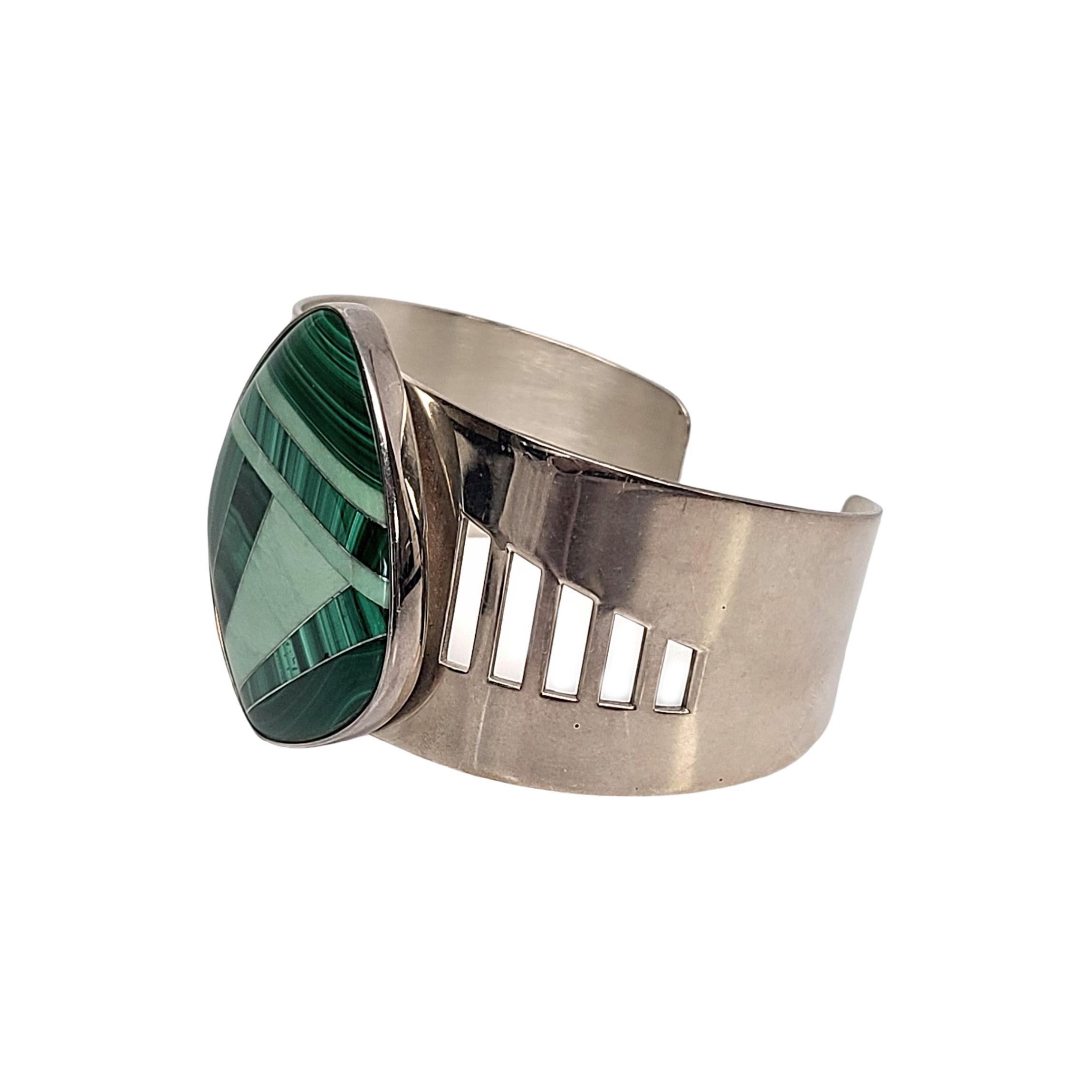 Breites Manschettenarmband aus Sterlingsilber und Malachit.

Mit einem breiten Band, das sich von vorne nach hinten verjüngt und mit rechteckigen Ausschnitten versehen ist. Großes Mosaik mit Malachit-Intarsien in der Mitte.

Maße ca. 5 3/4