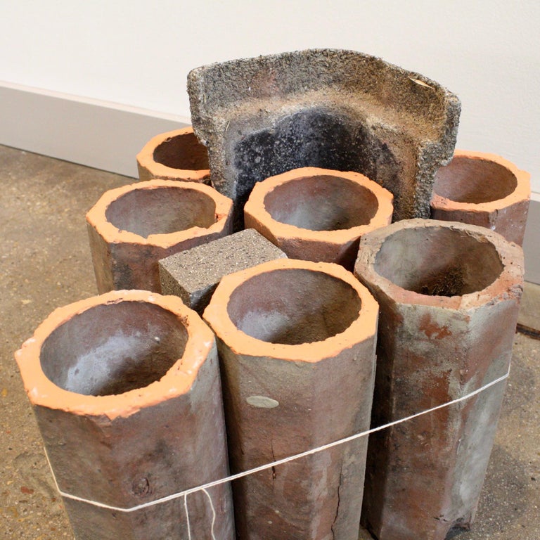 In this piece, seven octagonal terra-cotta bricks of cylindrical shape are on the floor in a V shape. Two concrete bricks are placed amongst these cylinders. All of these objects are bound together by a string that is tied around the objects.