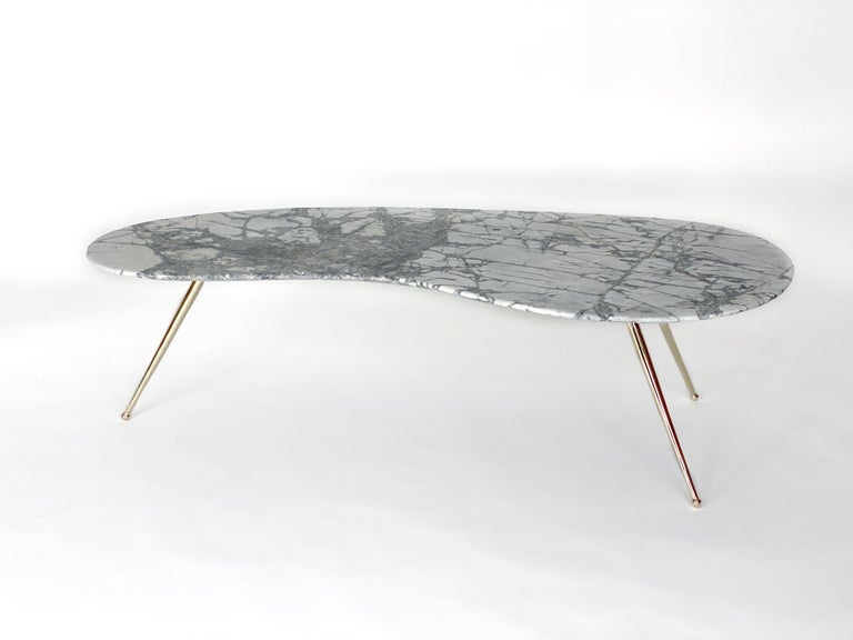 A 1960s inspiration. This elegant kidney shaped coffee table features a white Carrara marble top. The handcut slab, with it beveled edge, sits gracefully on three tapered brass legs. This sleek tripod base gives the sensation that the top is