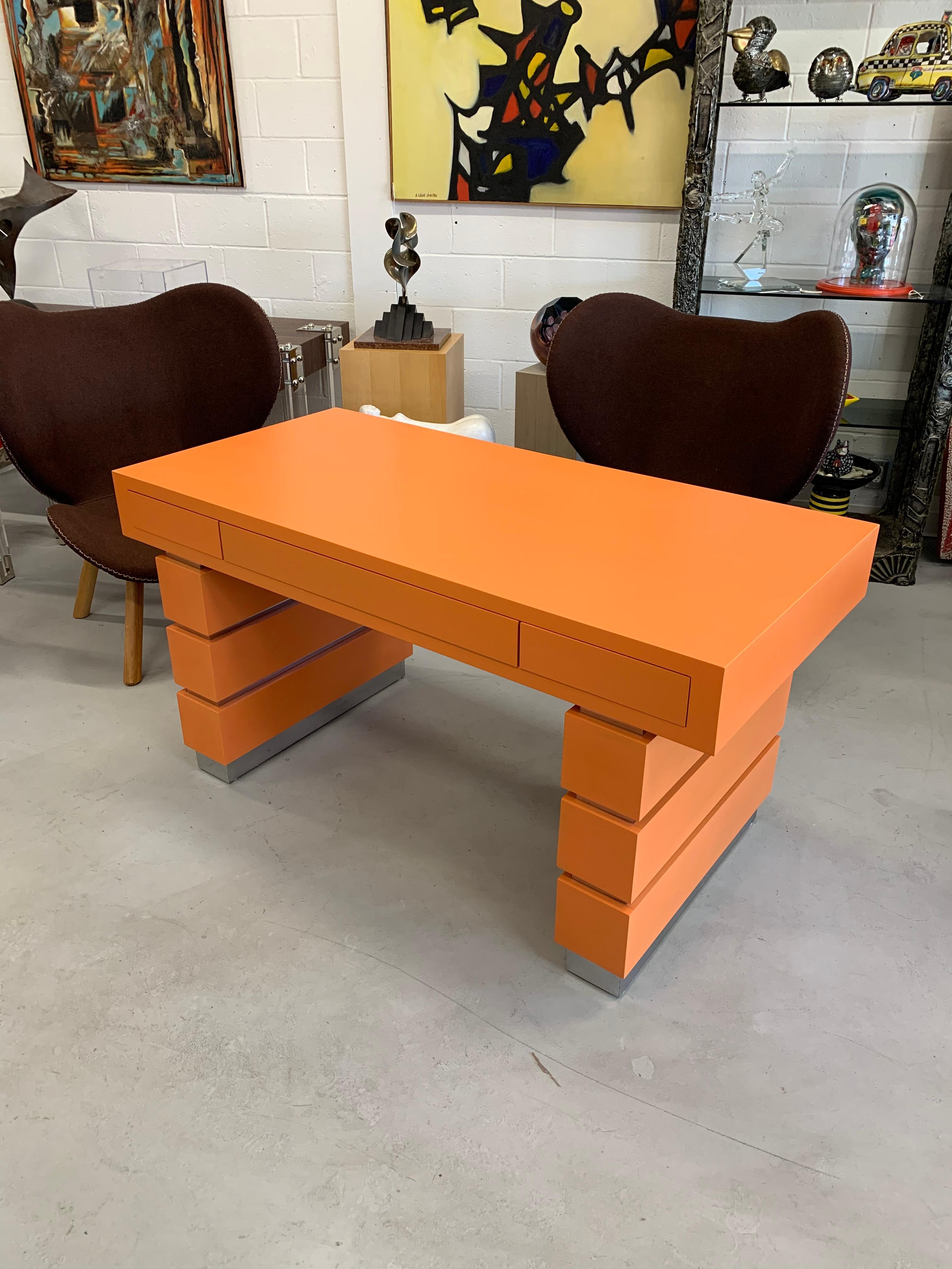 A beautiful custom orange and chrome banded desk with three drawers. It is finished on the back as well. It is approximately 57.75 by 27.75 inches in dimension and approximately 30.25 inches tall. Drawers have push in opening. This desk was produced