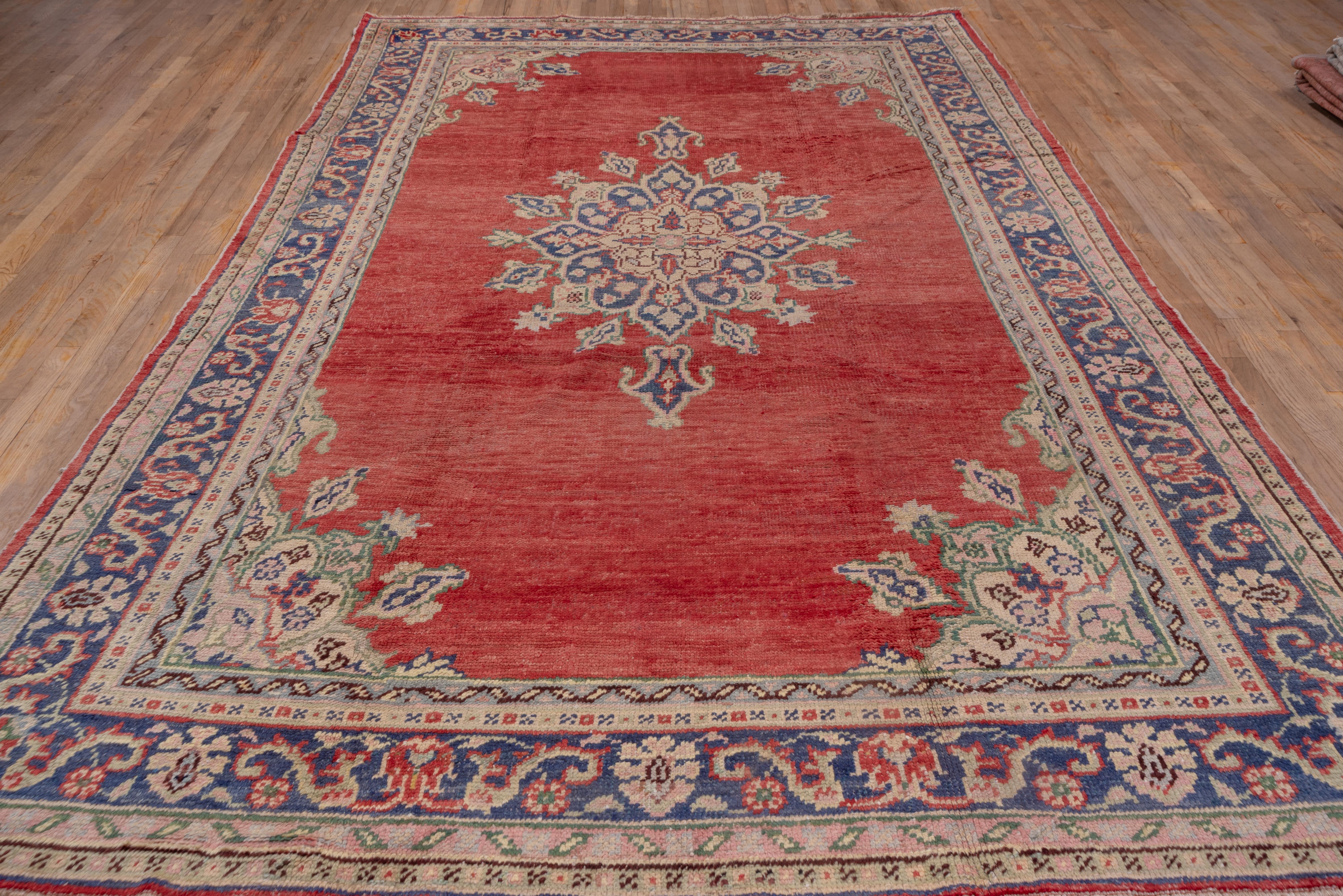 This west Anatolian town carpet shows a dramatic blue pendanted medallion with palmette periphery on an open red ground with sand corners and matching palmettes. The medium blue main border shows volute leaves and simple palmettes accented in pale