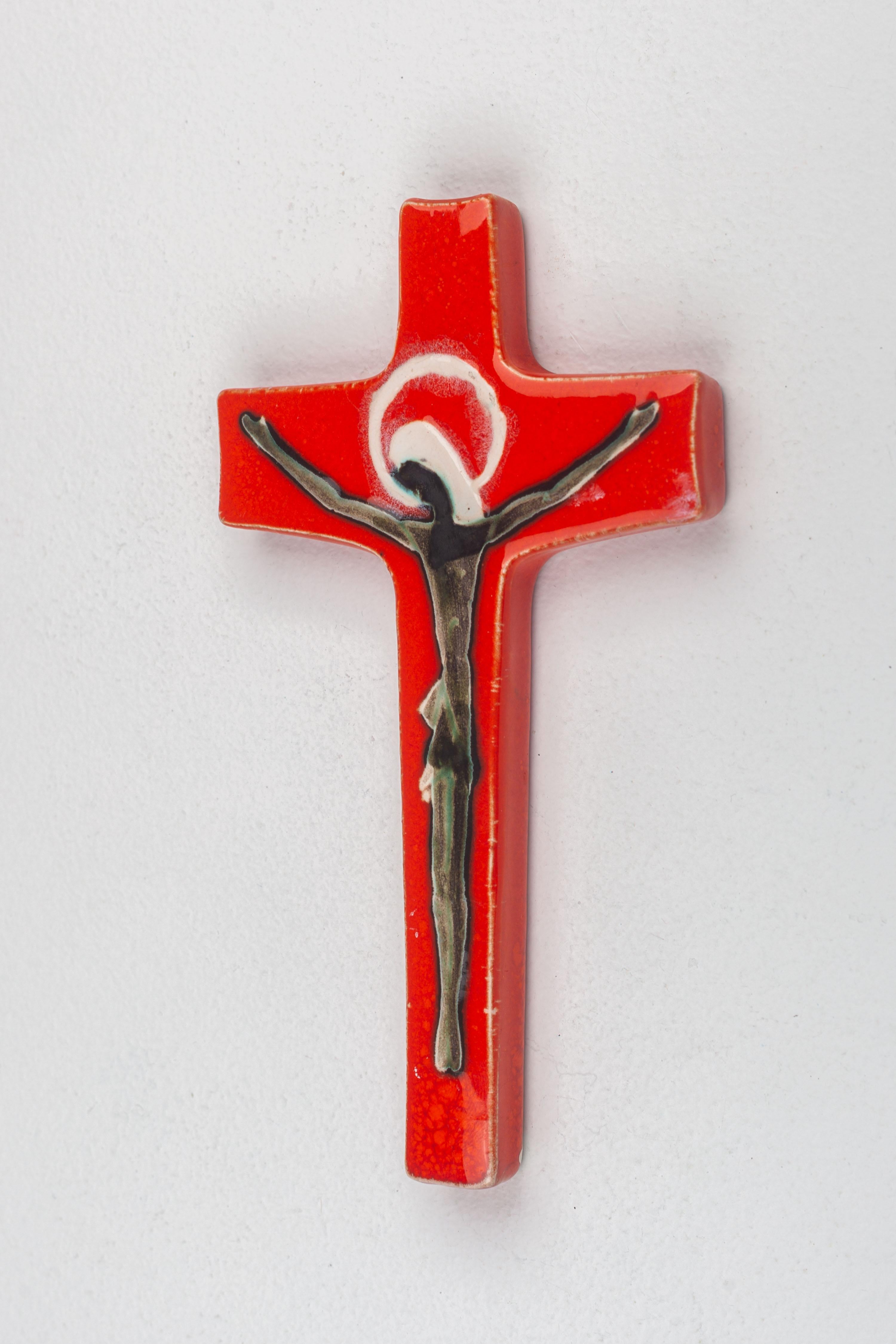 Ce crucifix en céramique est une représentation sublime de la poterie européenne de studio du milieu du siècle, réalisée à la main avec un soin méticuleux par un artisan qualifié. La croix est enveloppée d'une glaçure vermillon vibrante, qui lui