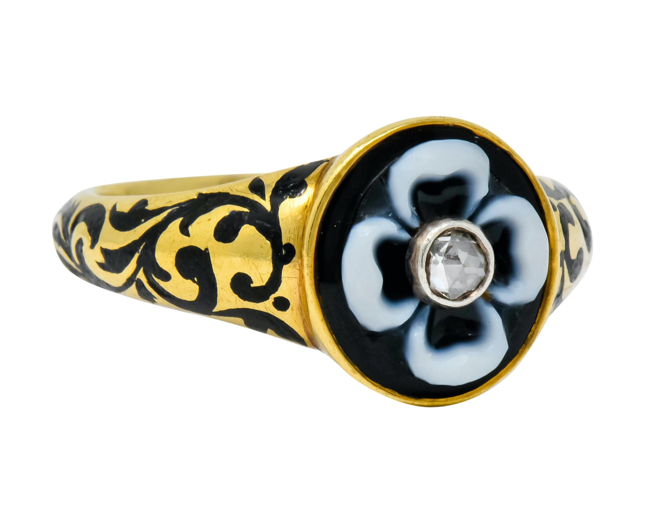 Centering a rose cut diamond, bezel set, in a carved onyx flower featuring white banding as its petals

Set in a matte gold mounting with scrolled enamel work fully around shank, black with some loss, consistent with age, wear, and use

With dated