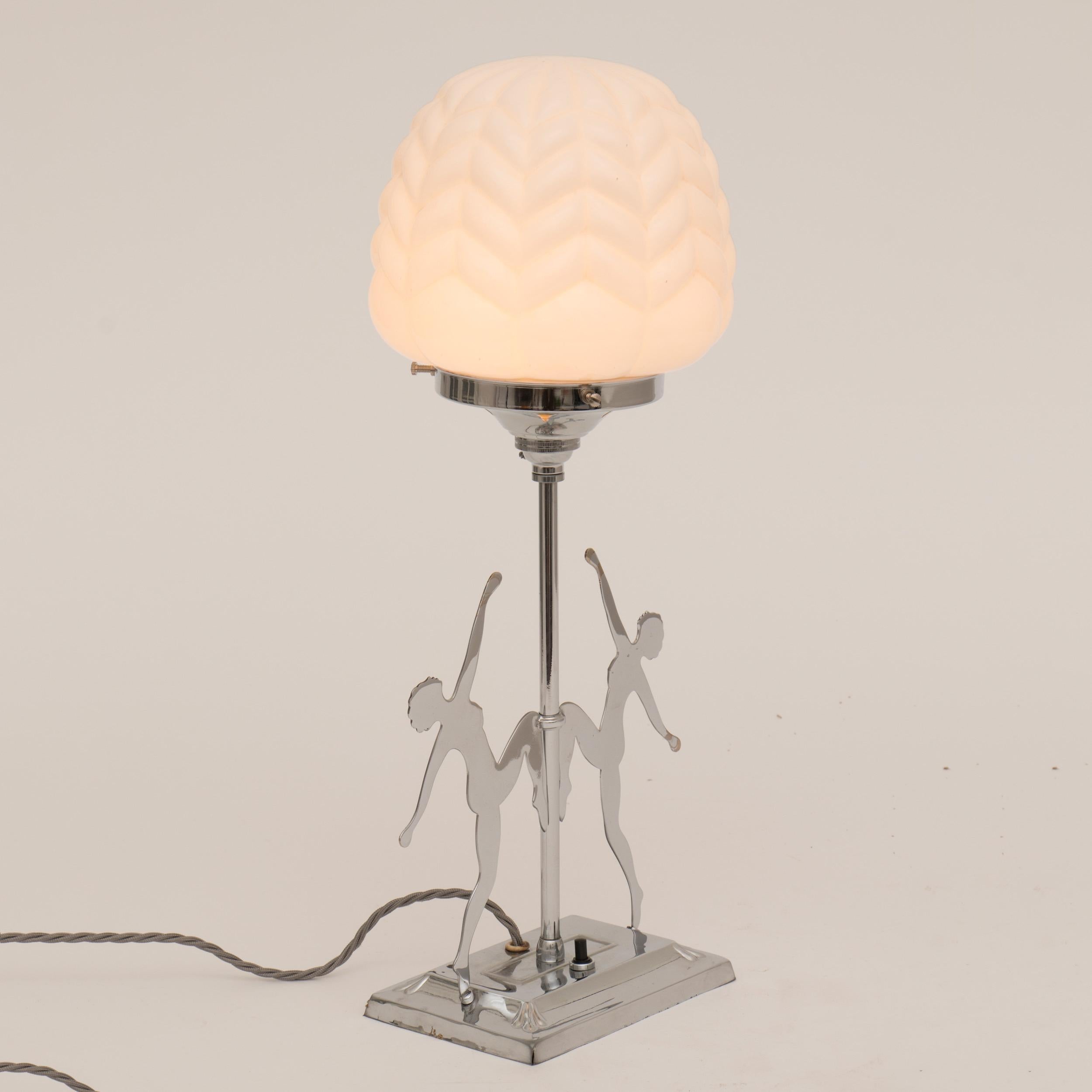 An Art Deco polished chrome table lamp with two dancing female silhouetted figures topped with a chevron pattered white glass shade
Measures: H 47cm, W 21cm, D 17cm
British, circa 1930.