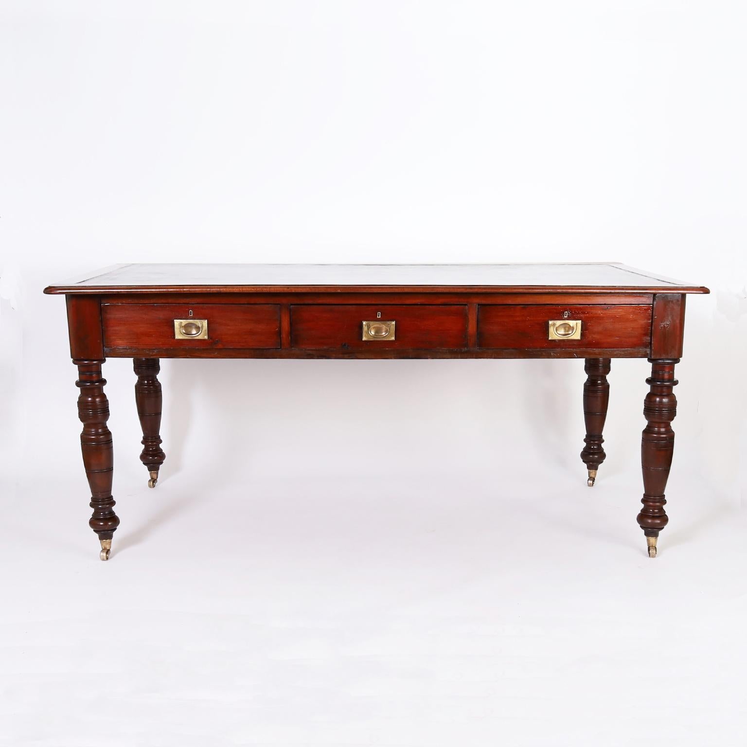 19th centur British colonial style desk or library table in mahogany with a variegated green brown tooled leather top, three drawers in the case with brass campaign hardware and turned legs with brass casters.