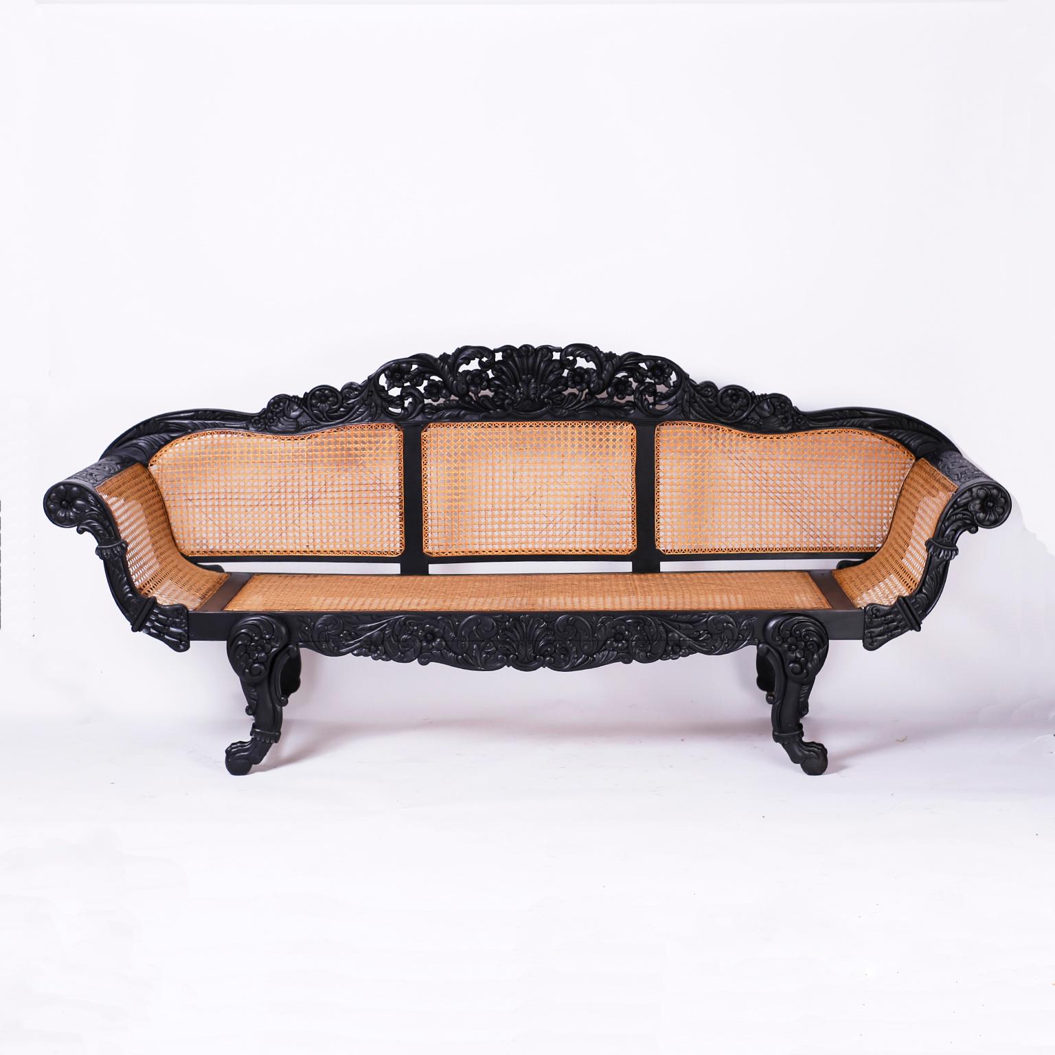 British colonial settee crafted in mahogany with an ebonized finish. Featuring an elaborately carved floral crest with center sea shell, hand caned seat and back, floral carved arms and skirt, dramatic form and claw and ball feet.