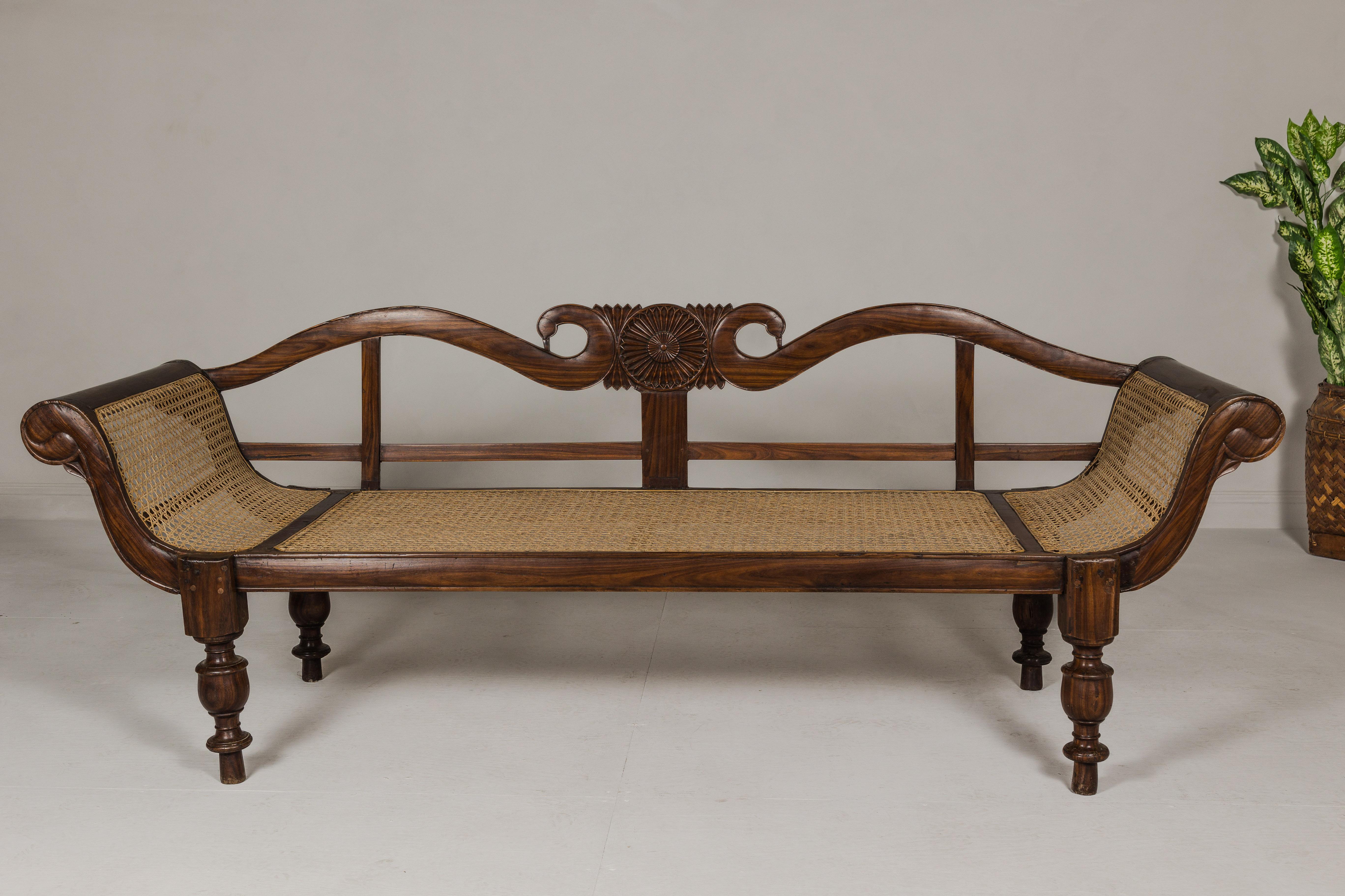A British Colonial carved wooden settee with swan neck back and out-scrolling arms, cane seat and turned legs. This British Colonial carved wooden settee, originating from antique South-East Asia, is a striking blend of elegance and colonial design
