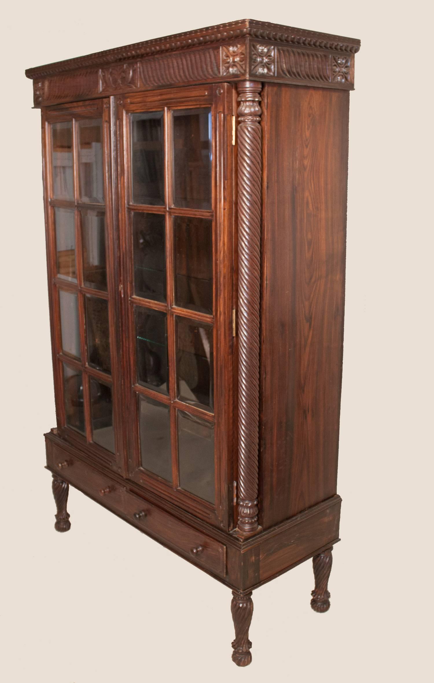 An intricately hand-carved and gadrooned rosewood cabinet with paned, beveled glass doors, circa 1960. This restored British Colonial style piece is crafted in two parts, with a locking cabinet sitting atop a two-drawer stand on shapely legs. Three