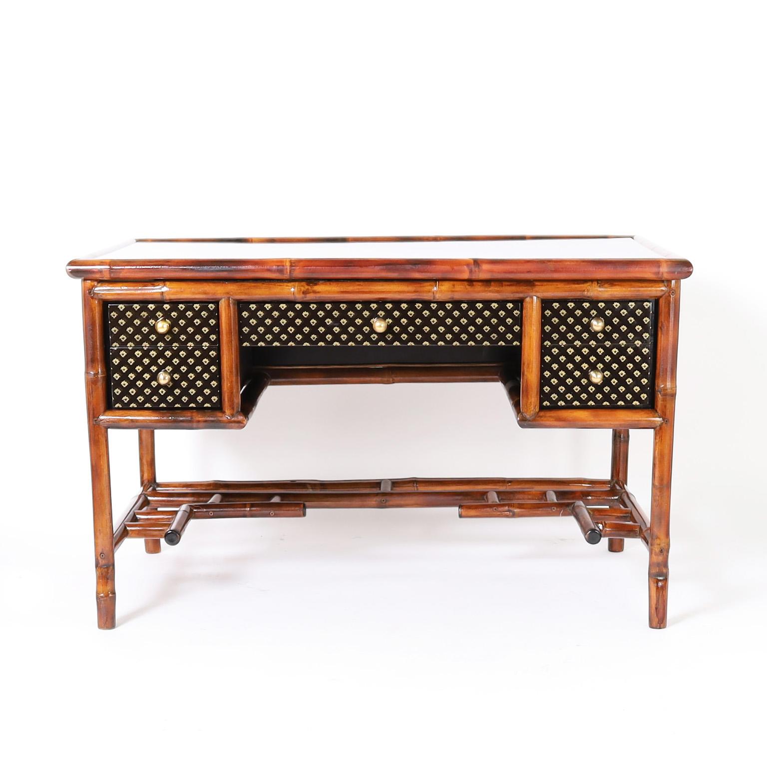 Mid century desk crafted in bamboo and featuring lacquered panels on the top under glass, sides, back and front having five drawers with brass hardware and Chinese Chippendale style stretcher below.