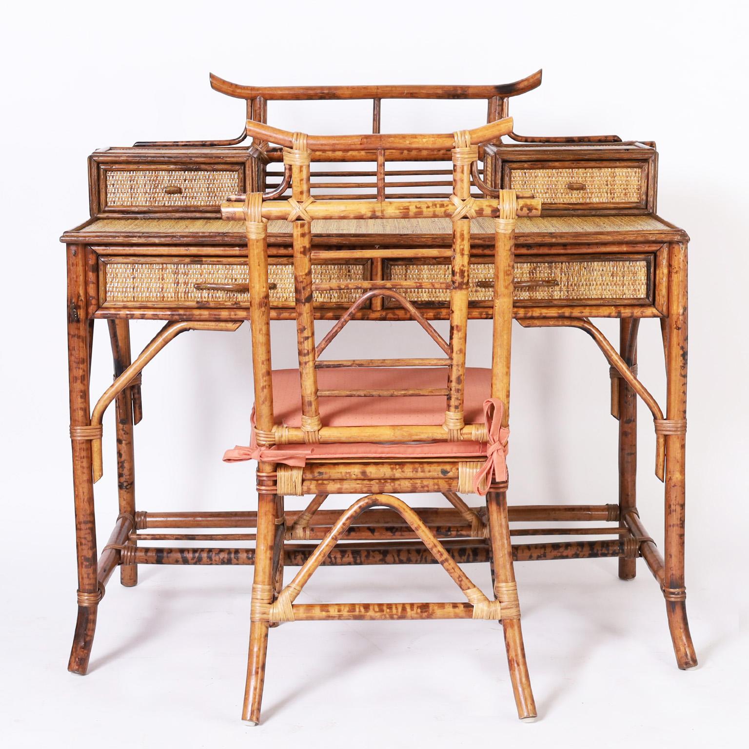 Midcentury British colonial style desk and chair crafted with a burnt bamboo frame having pagoda form galleries. The desk has two glove boxes, grasscloth panels on a case with three drawers, Asian style brackets and splayed feet. 

Chair measures