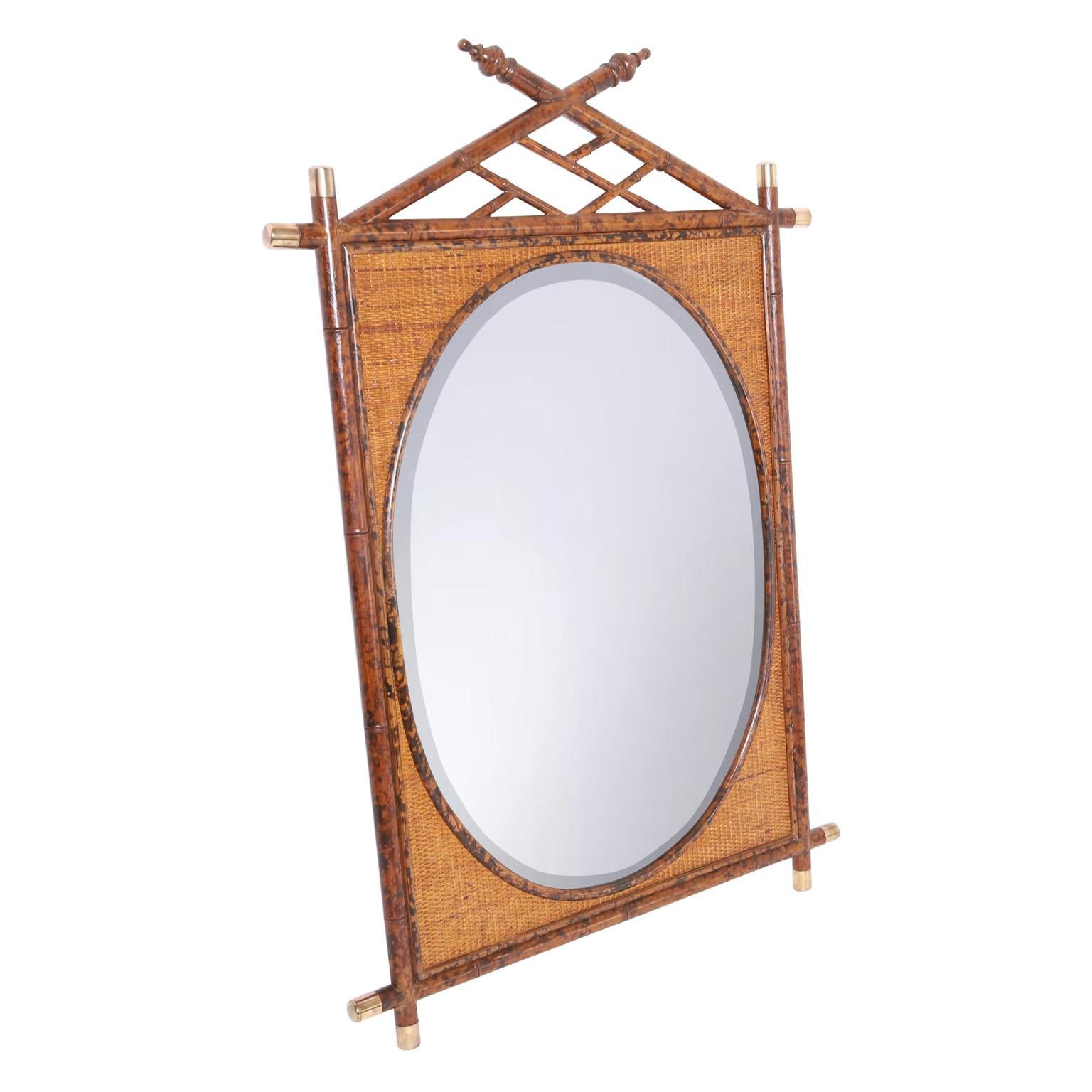 British colonial style wall mirror having a rectangular frame crafted in faux burnt bamboo with brass caps, a chippendale style crest, and grasscloth panels around an oval beveled looking glass.