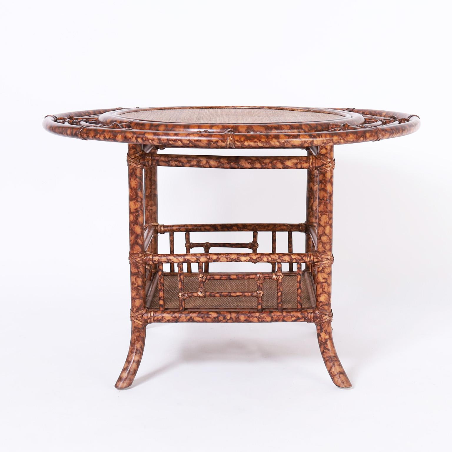 British colonial style coffee table crafted in faux bamboo with a faux tortoise finish having a round top with a grasscloth panel in the middle. The base has a lower tier and splayed legs.