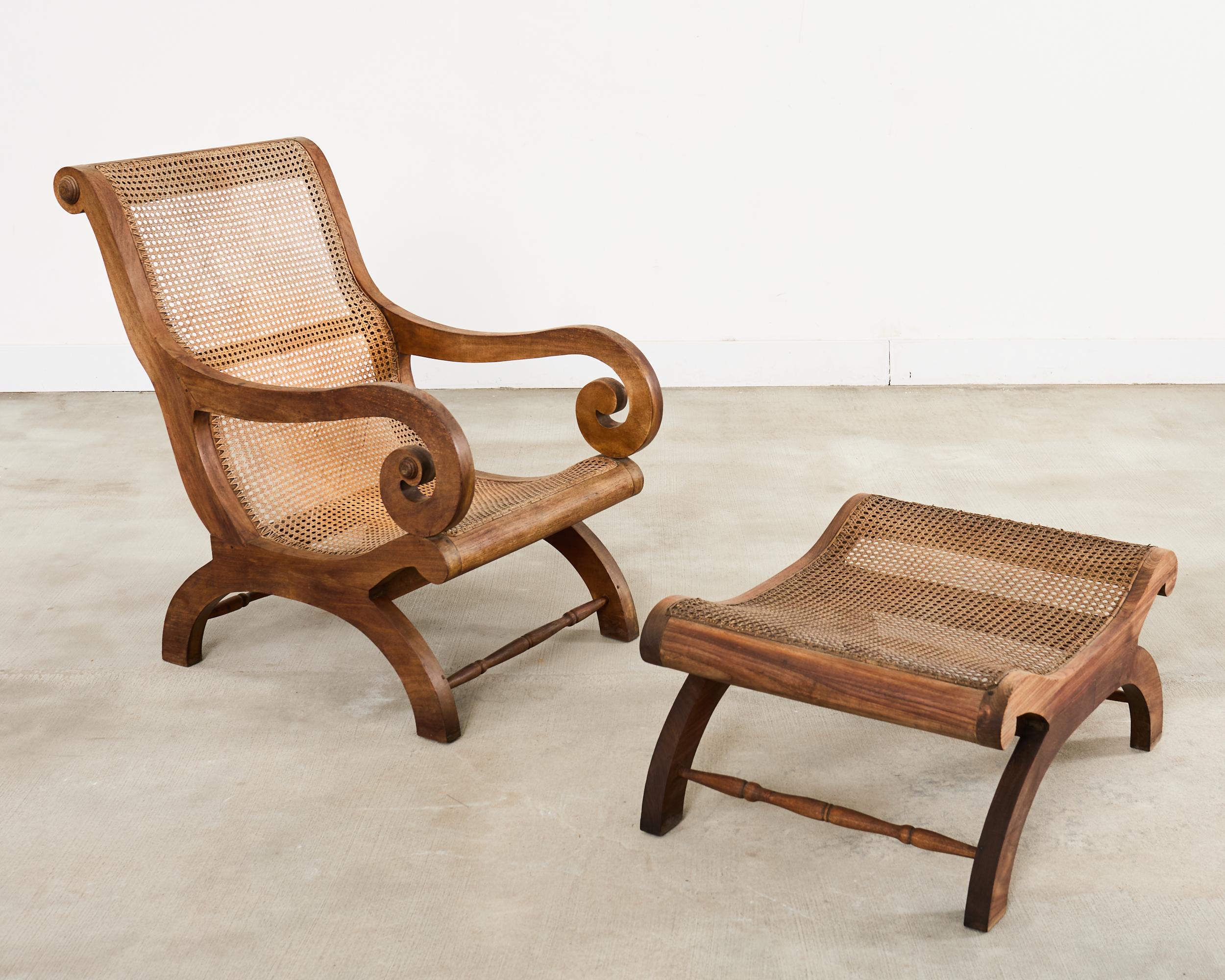 Splendida poltrona anglo-indiana da salotto con pouf abbinato, realizzata in stile Grand British Colonial. La sedia presenta una struttura in teak graziosamente intagliata con ampi braccioli a voluta. Il sedile e lo schienale sono rivestiti di canne