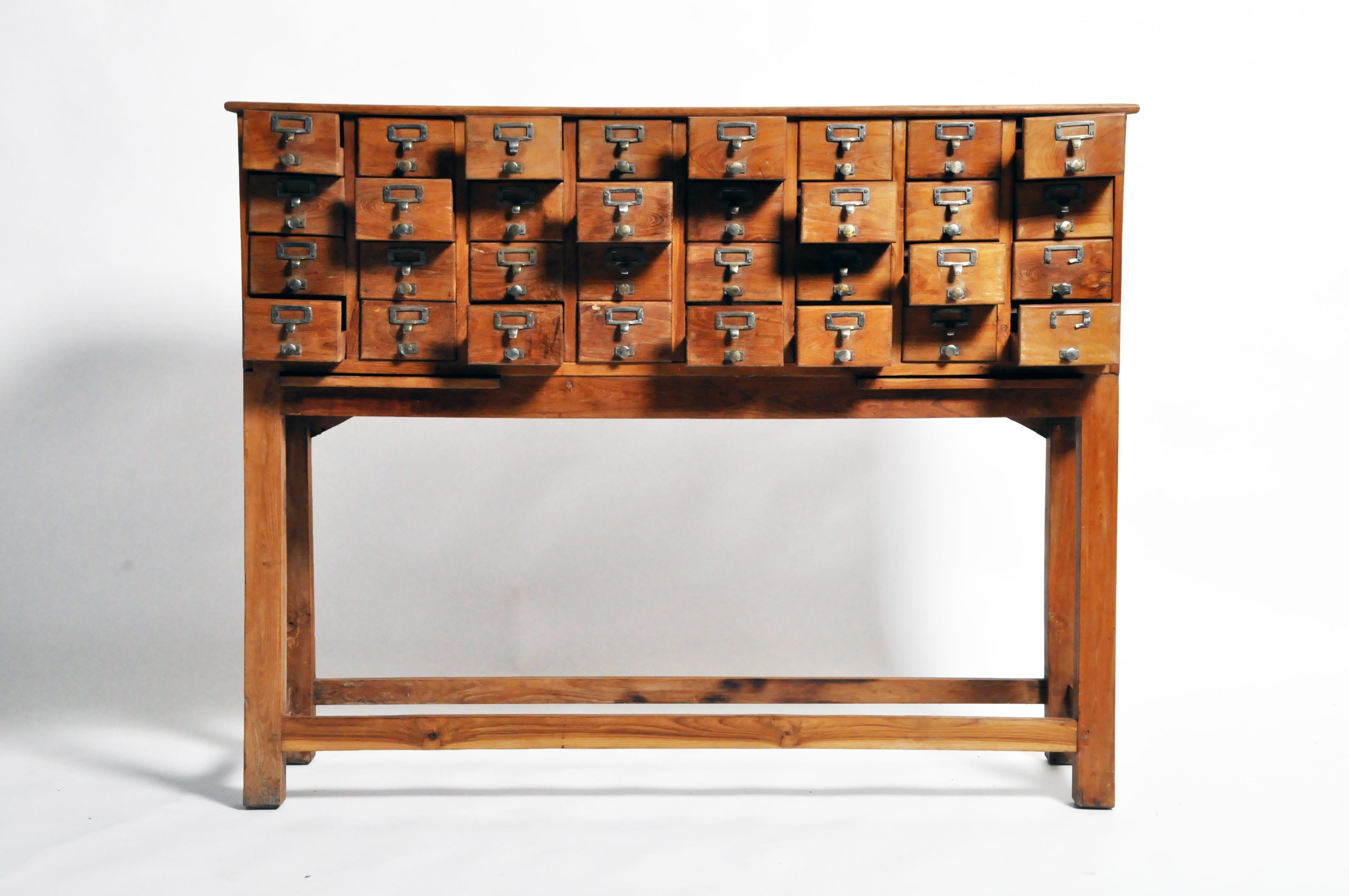 This impressive chest features 32 drawers reminiscent of a library card catalog and two retractable shelves. Made of teakwood, the piece has a lovely golden hue and visible grain. At nearly five feet in height, the chest is functional and a lovely