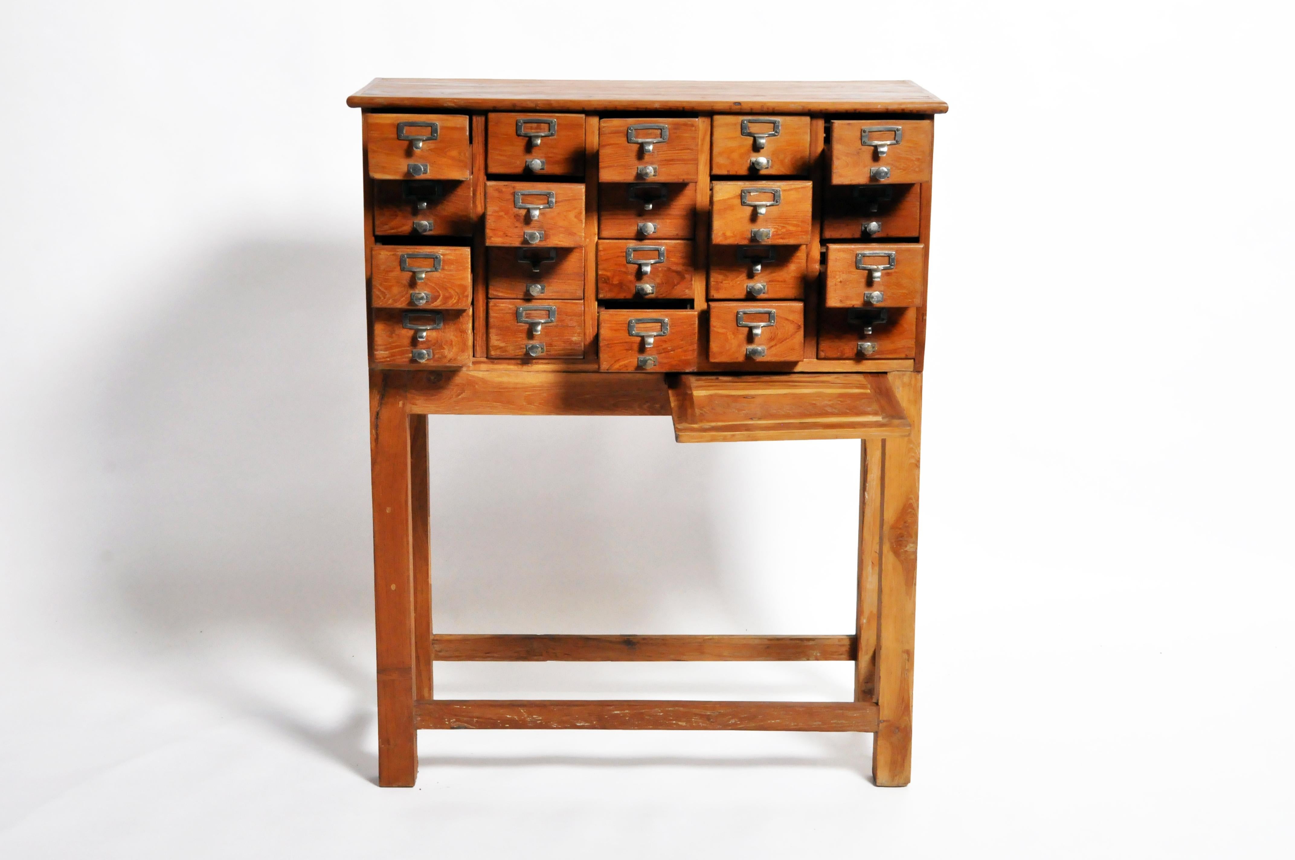 This impressive chest features 20 drawers reminiscent of a library card catalog and two retractable shelves. Made of teak wood, the piece has a lovely golden hue and visible grain. At nearly five feet in height, the chest is functional and a lovely