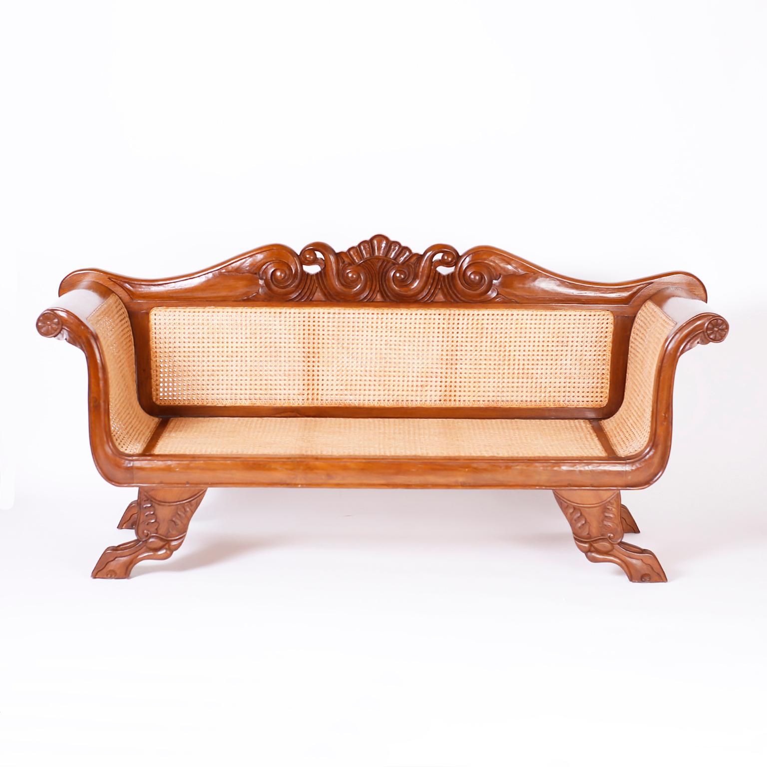 19th century William IV Caribbean or West Indian settee hand crafted in mahogany with an ocean inspired center carving, rolled arms, caned seat back and sides, pegged construction, and carved winged animal paw feet.