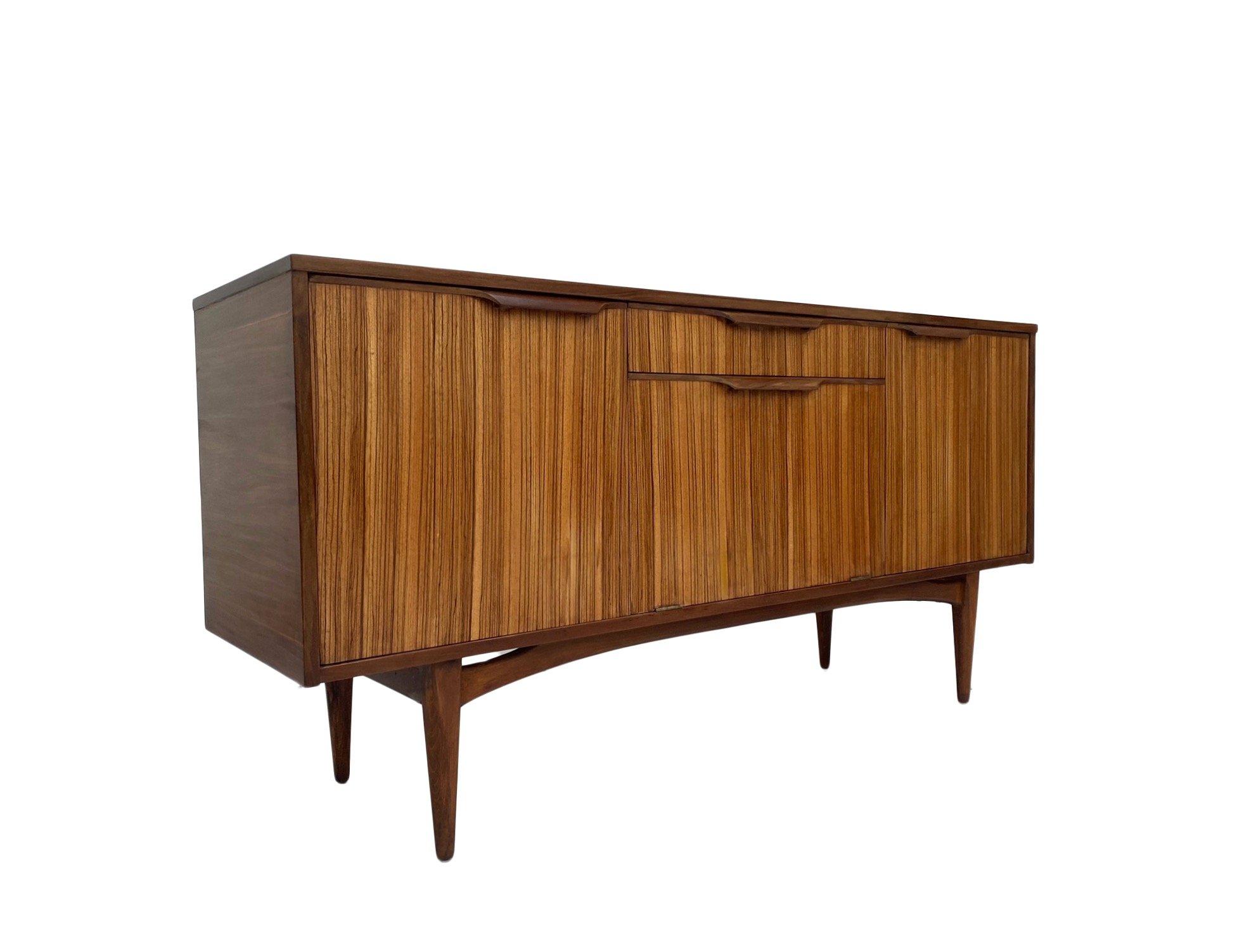 A beautiful British teak sideboard designed by Elliots of Newbury, this would make a stylish addition to any living or work area.

The cabinet has a cupboard with shelf to either end and a drop down cupboard and drawer to the centre.

The sideboard