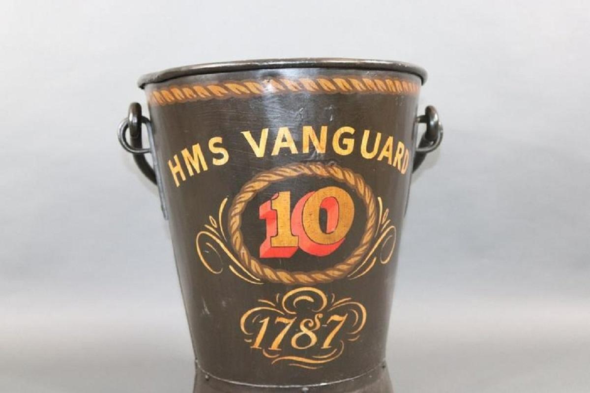 English Navy deck bucket circa 1930 with later decoration for HMS VANGUARD and dated 1787. The black painted bucket with decorative rope border is numbered 10. 12T 14W x 17D.