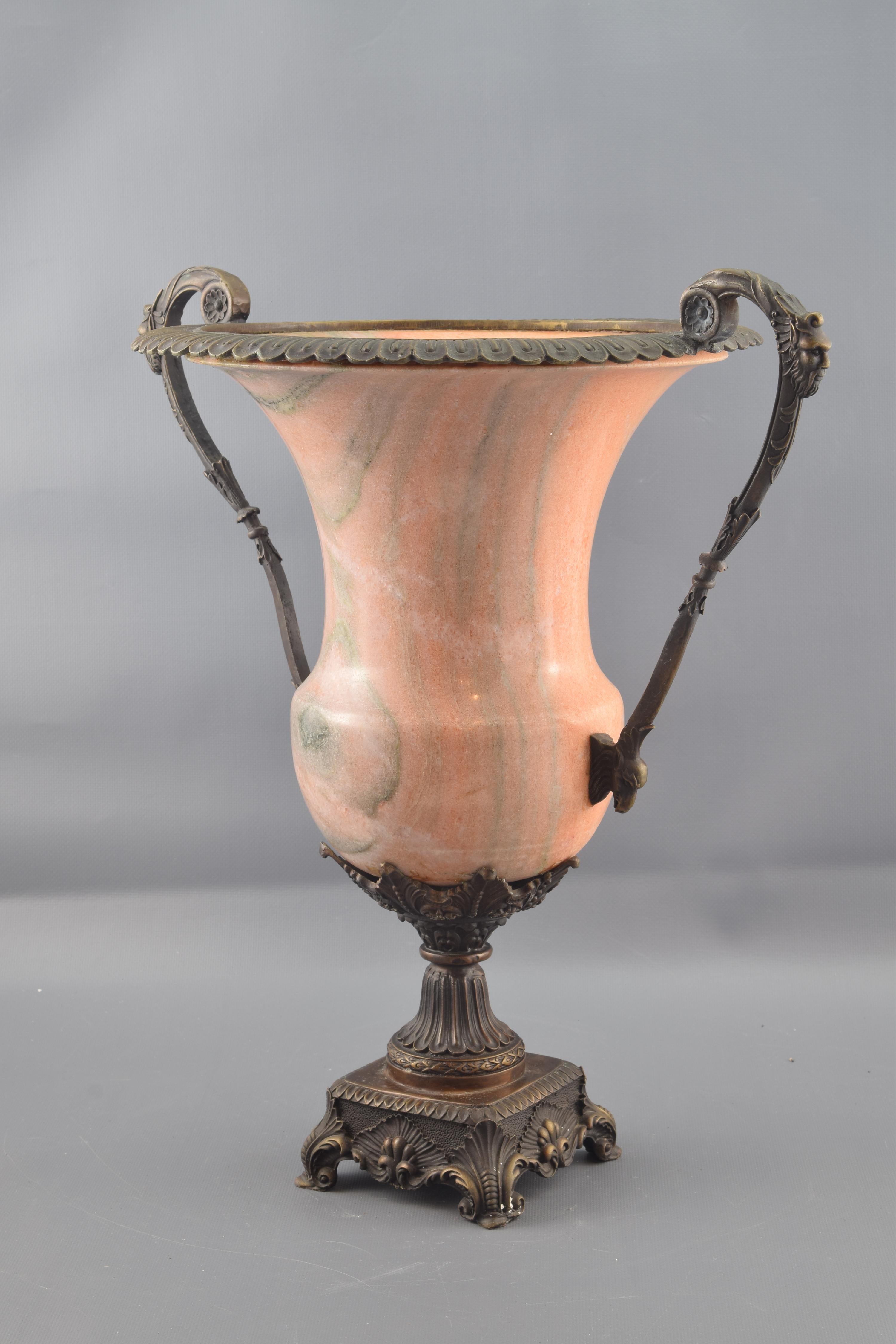 On a square base supported by scrolls and decorated with vegetal motifs, the cup is raised, formed by a foot and handles with masks and leaves in bronze and a container in pink tone alabaster. The classical inspiration of the work is clear both by