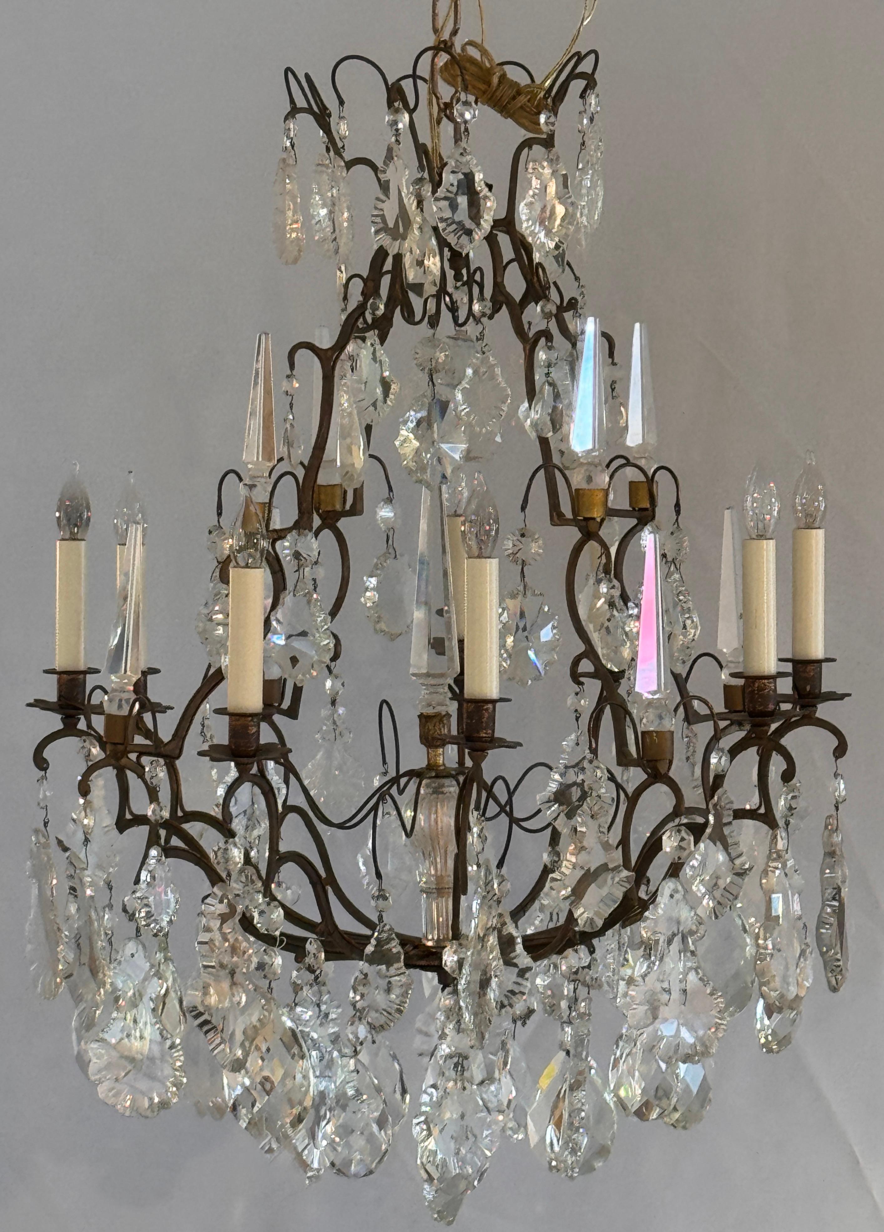 Lampadario a dodici luci con struttura in bronzo e ornamenti in cristallo riccamente dettagliati, tra cui grandi gocce sfaccettate, rosette tagliate e alti prismi in stile obelisco. Gli eleganti bracci curvilinei e la struttura a gradini offrono una
