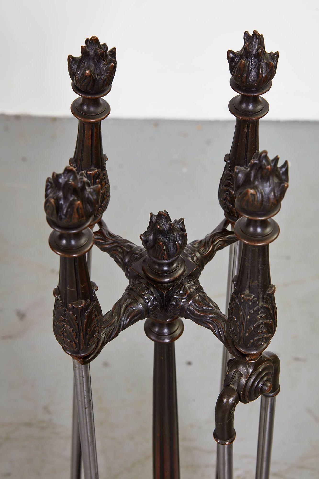 A set of four firetools with decorative flame bronze handles and polished steel shafts, consisting of a shovel, tongs, hooked poker and fireplace brush of real horsehair, on a finely cast tall bronze stand with central column topped with flame