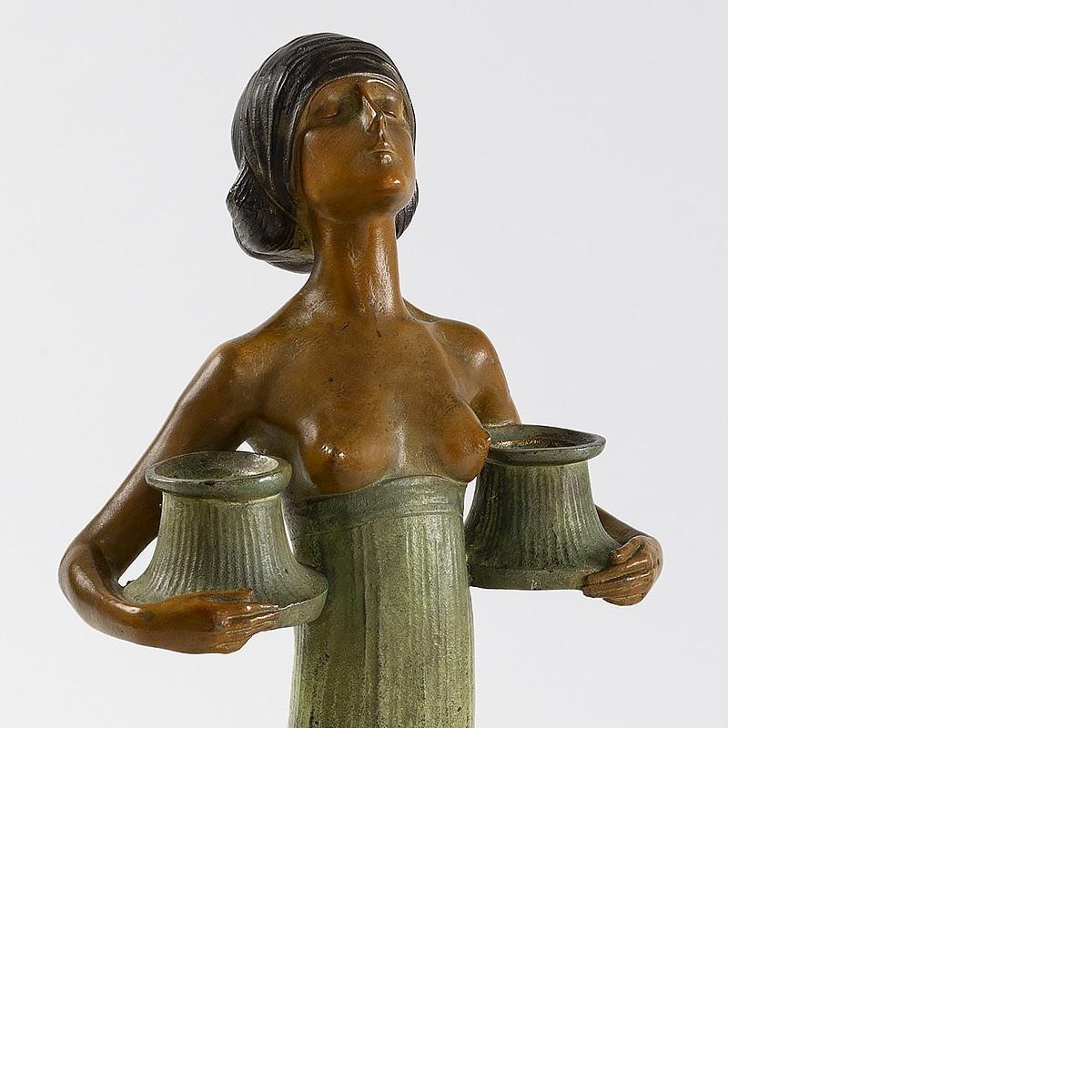 A polychrome bronze Art Nouveau figural candelabrum by Gustav Gurschner. A young woman with brown hair, dressed in a long flowing green skirt, with her breasts exposed, holds a seed capsule in each arm, into which a candle can be inserted. Gurschner