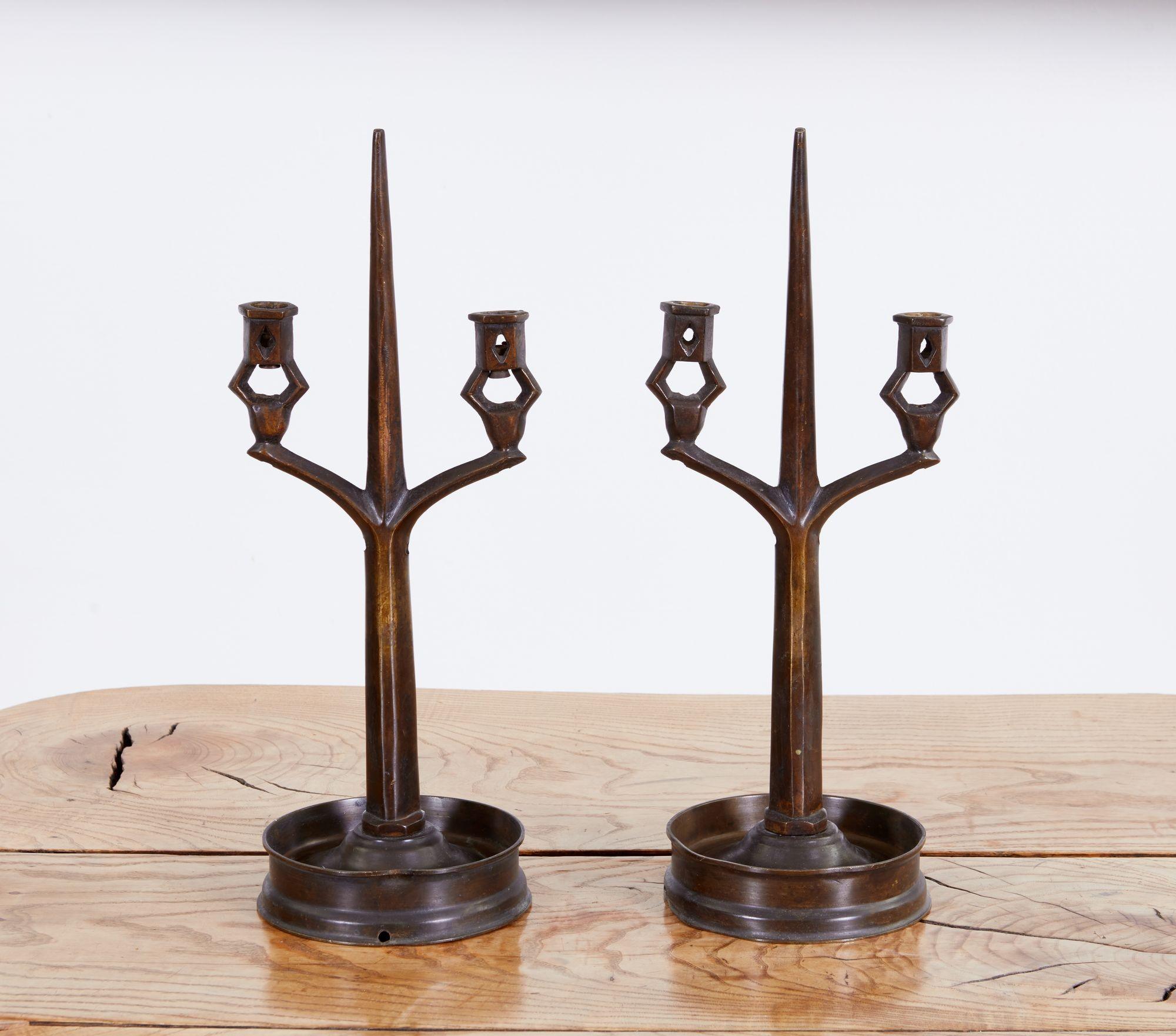 A pair of Arts and Crafts period two light candlesticks made of heavy bronze. Central tapered octagonal pricket surmounted by twin open candle sockets over turned base with deep drip pan, in a stylized gothic manner. Good rich glowing chocolate
