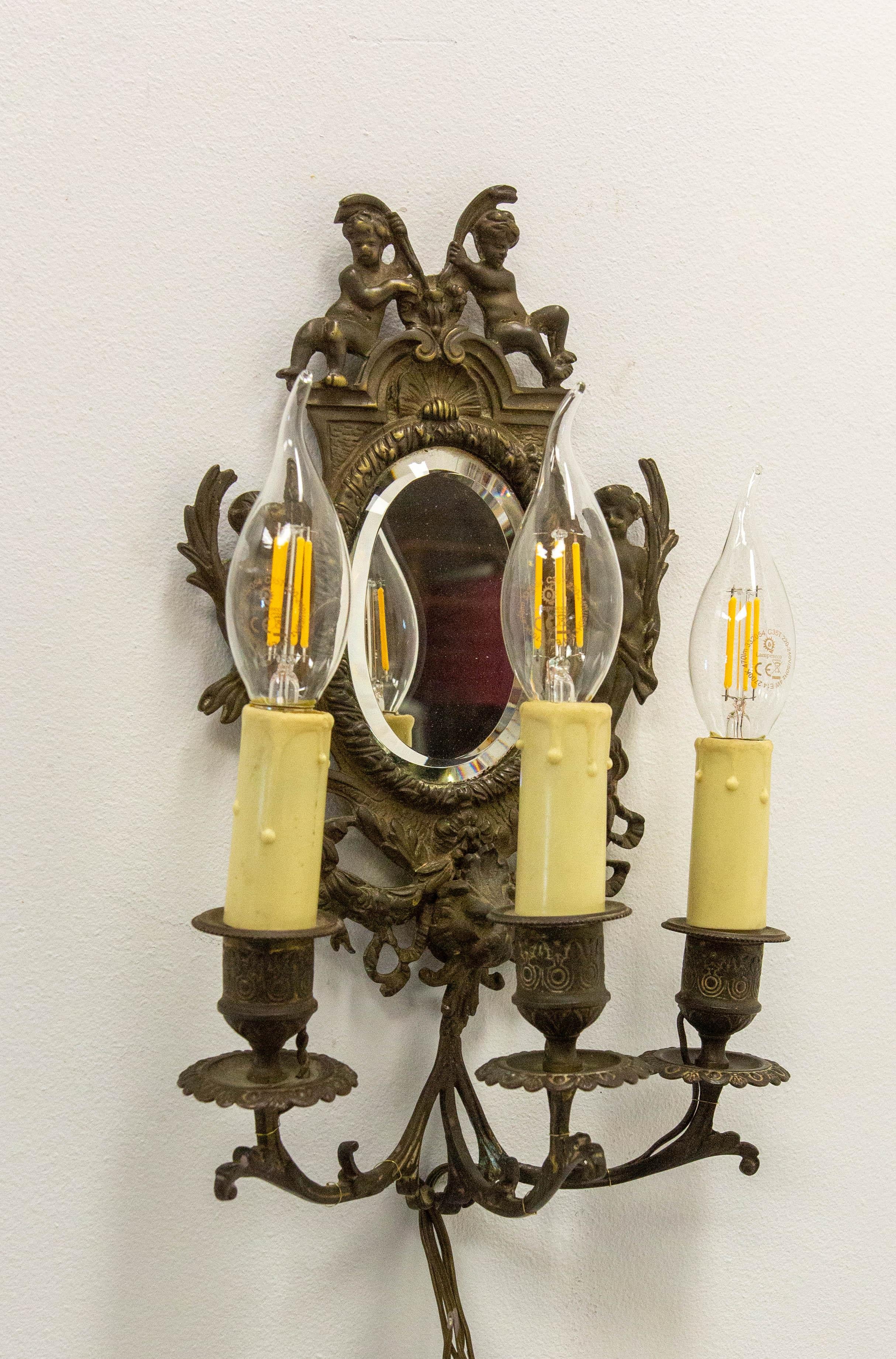 Aplique de bronce francés con espejo biselado ovalado y tres luces.
El espejo está rodeado por cuatro querubines y vegetación de estilo muy clásico. También está rematada por una concha, otro ornamento típico de Luis XVI.
El espejo biselado es el