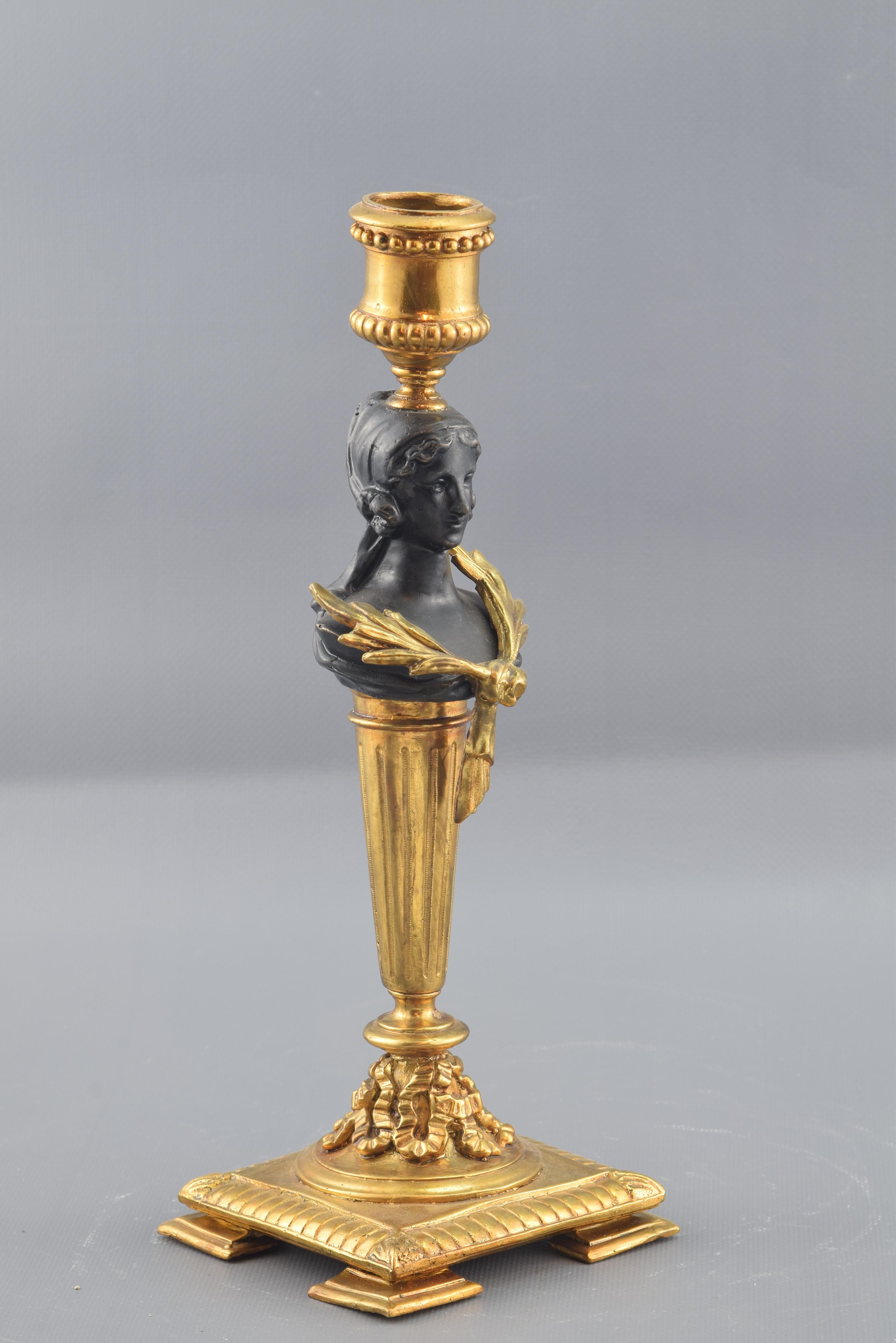 Patinated and gilded bronze.
On a square base decorated with eggs, stands an inverted capital with vegetal motifs. The foot of the candlestick is formed by a stipe of smooth circular shaft, on which sits an Hermes with laurels on the chest, which