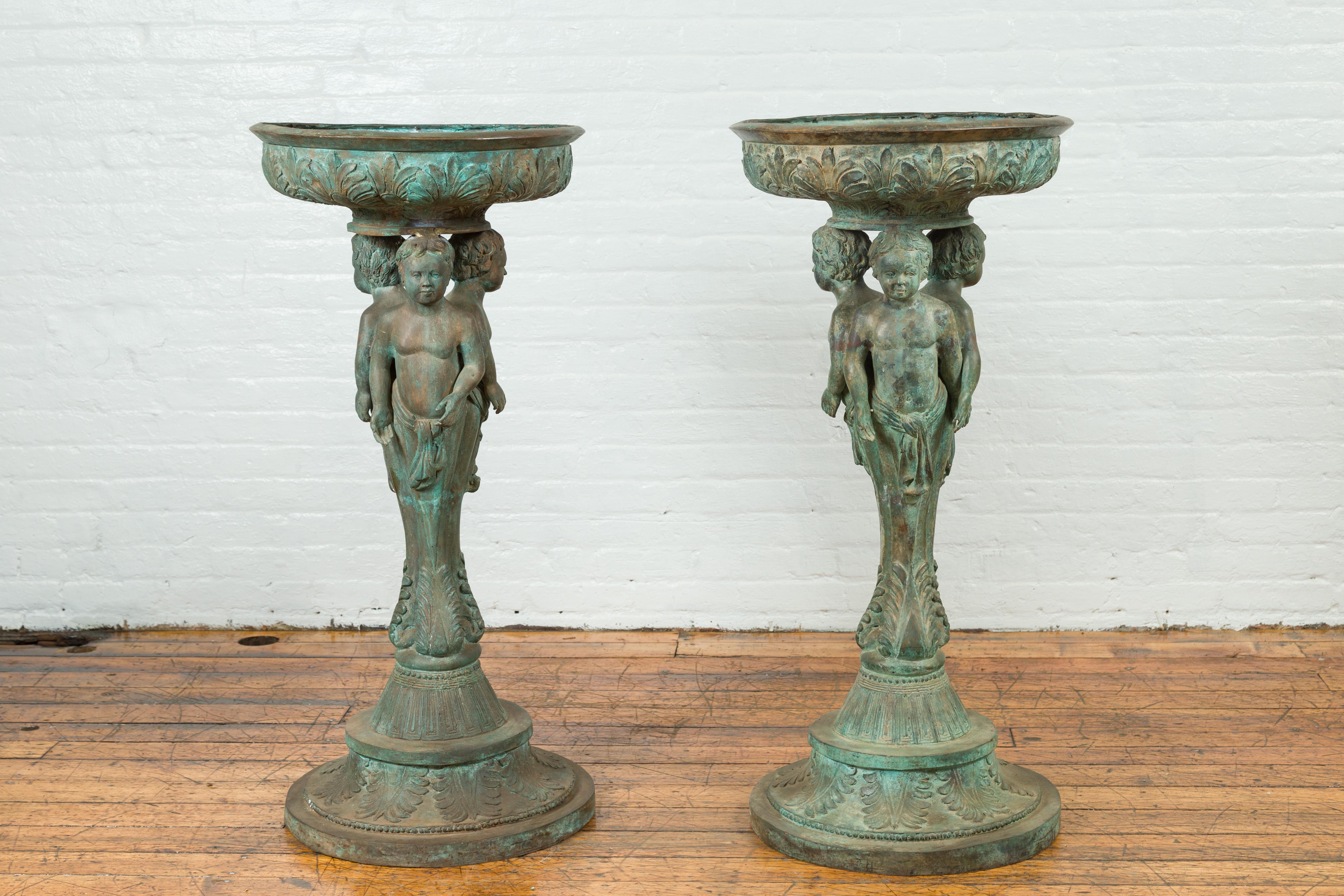 This pair of bronze lost-wax classical planter urns from the 20th century feature three cherub figures that merge together to create the base of these bronze pedestals. The three cherub boys' legs blend together to a narrow point where the base