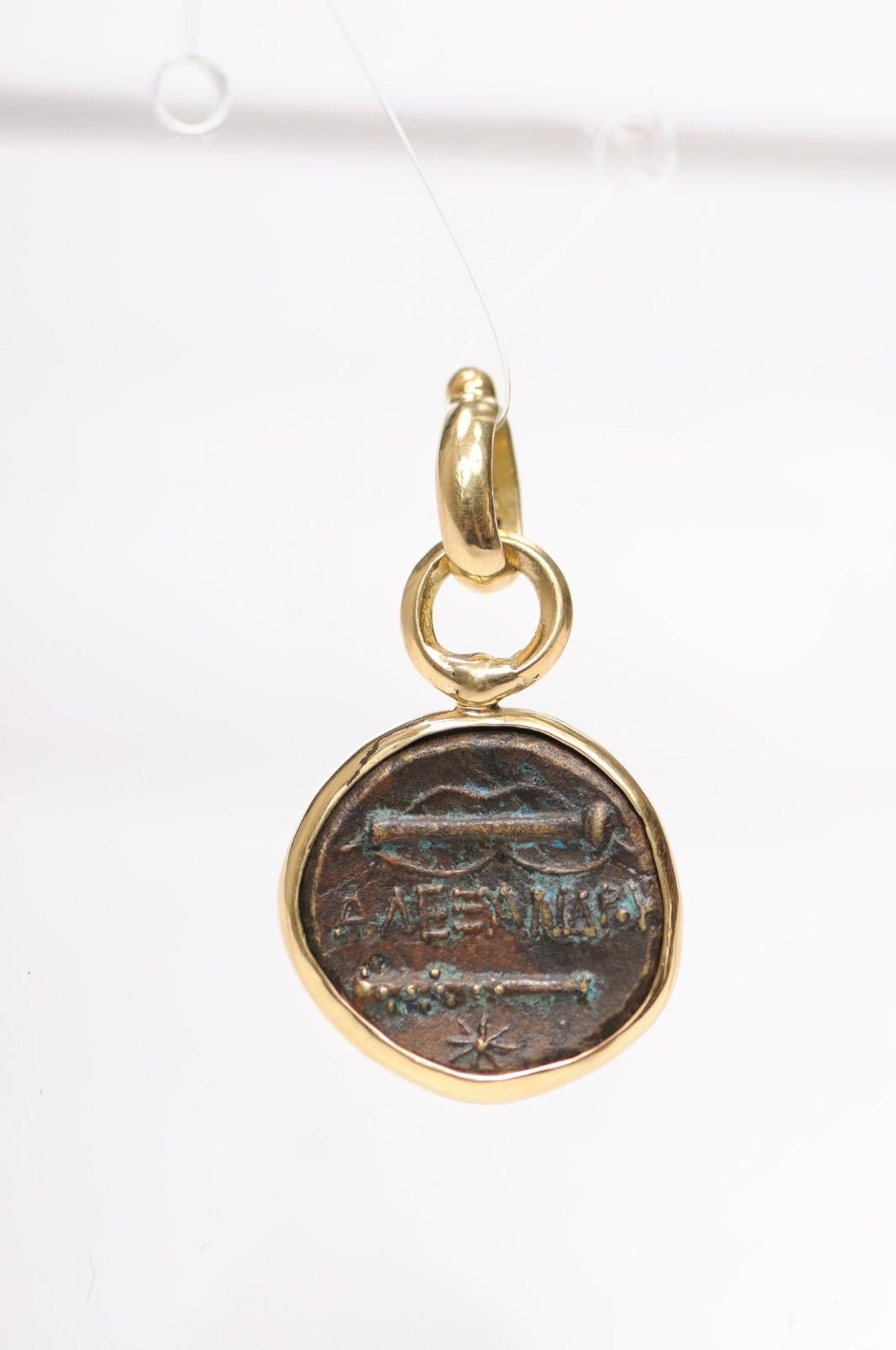 Un ciondolo a forma di moneta in bronzo rifinito in oro 18 carati. Questa moneta proviene dal Regno di Macedonia, circa 331-323 a.C.. Sul dritto, la testa di Eracle rivolta a destra, con indosso una pelle di leone. Sul retro, ci sono delle mazze con