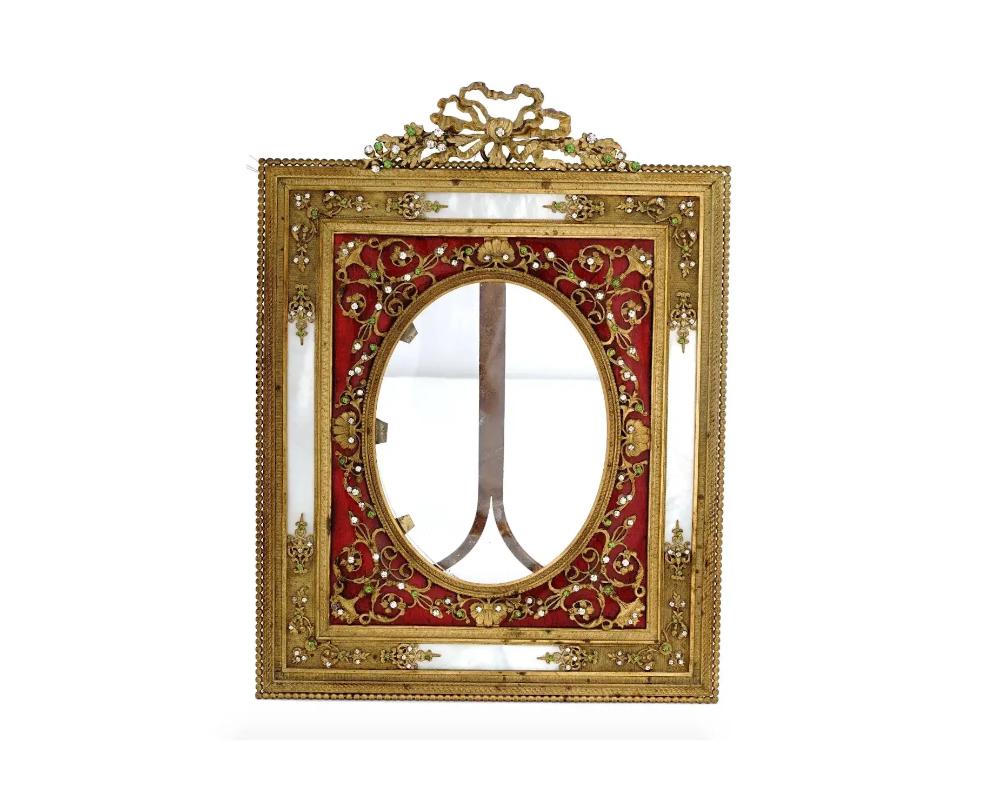 An antique French gilt bronze picture frame decorated with applied Rococo style adornments depicting floral motifs set with green and white rhinestones. The frame is also decorated with red guilloche enamel and pearlescent white inserts. The