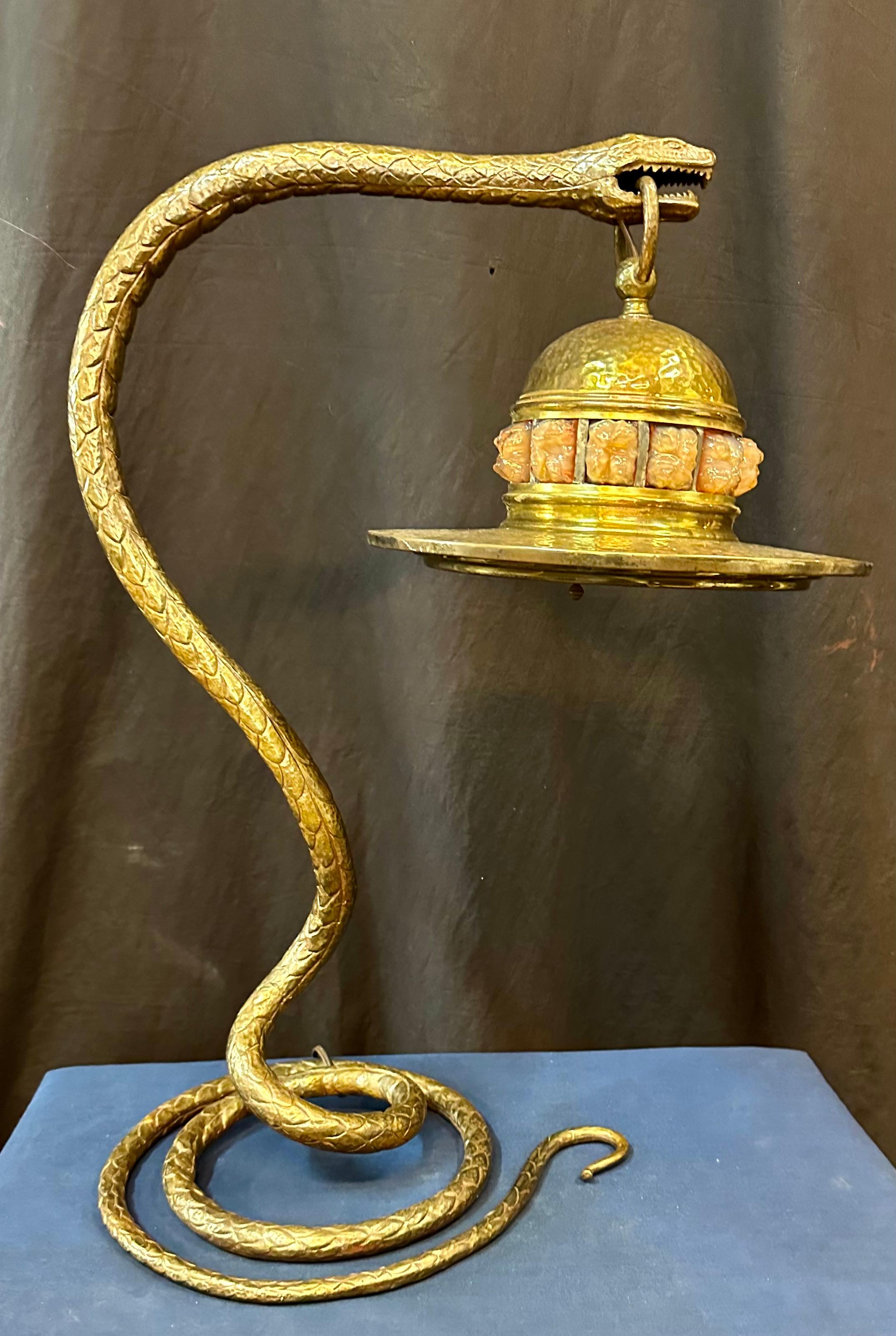 This vintage golden bronze French table lamp dates from the early 20th century. A realistically rendered sculpted bronze coiled snake is holding the top ring of a hand hammered lamp shade in its mouth. This shade is designed with a bold band of