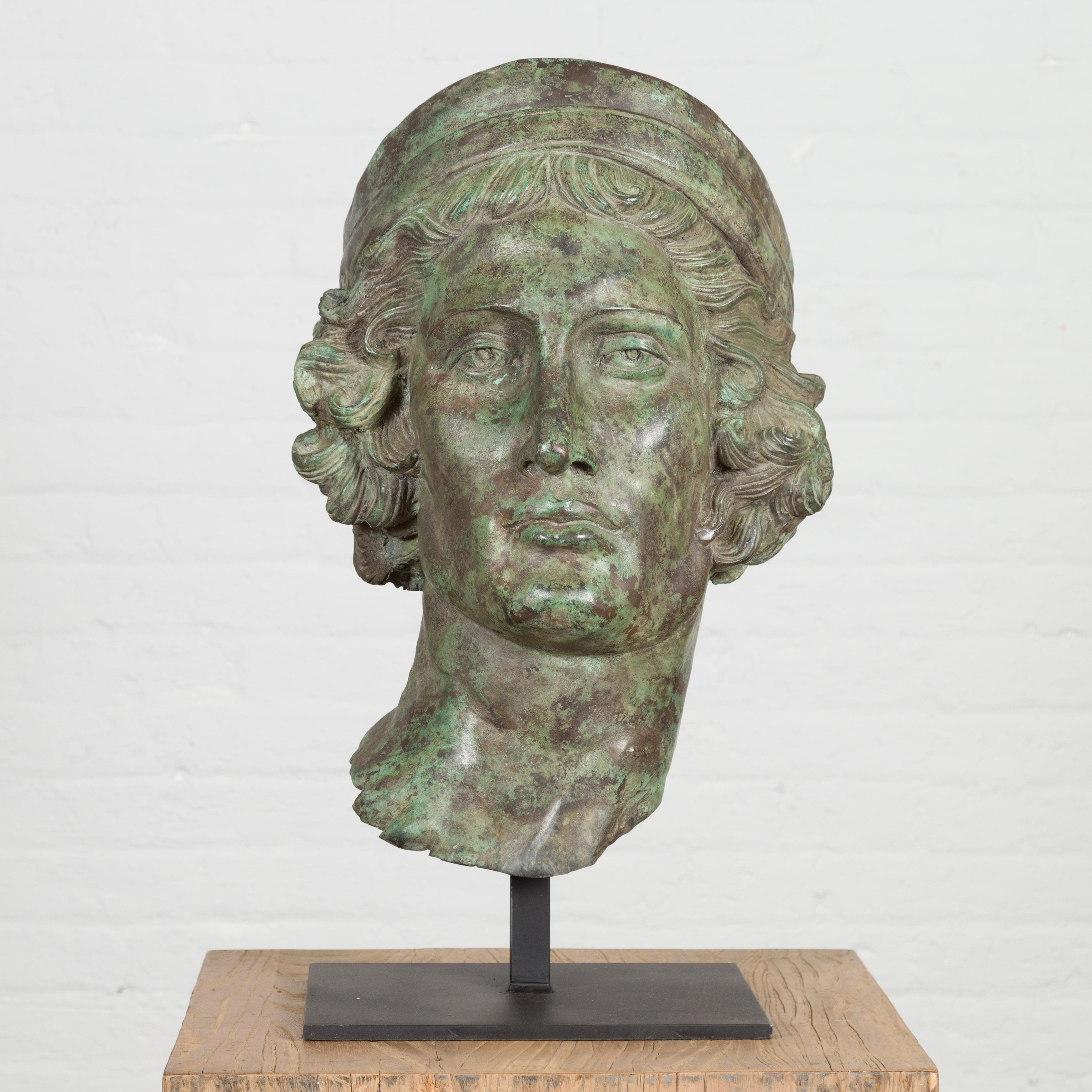 Sculpture de tête de style gréco-romain en bronze coulé à la cire perdue représentant un jeune avec un bandeau en patine vert-de-gris, monté sur une base personnalisée de couleur noire. Créée selon la technique traditionnelle de la cire perdue qui