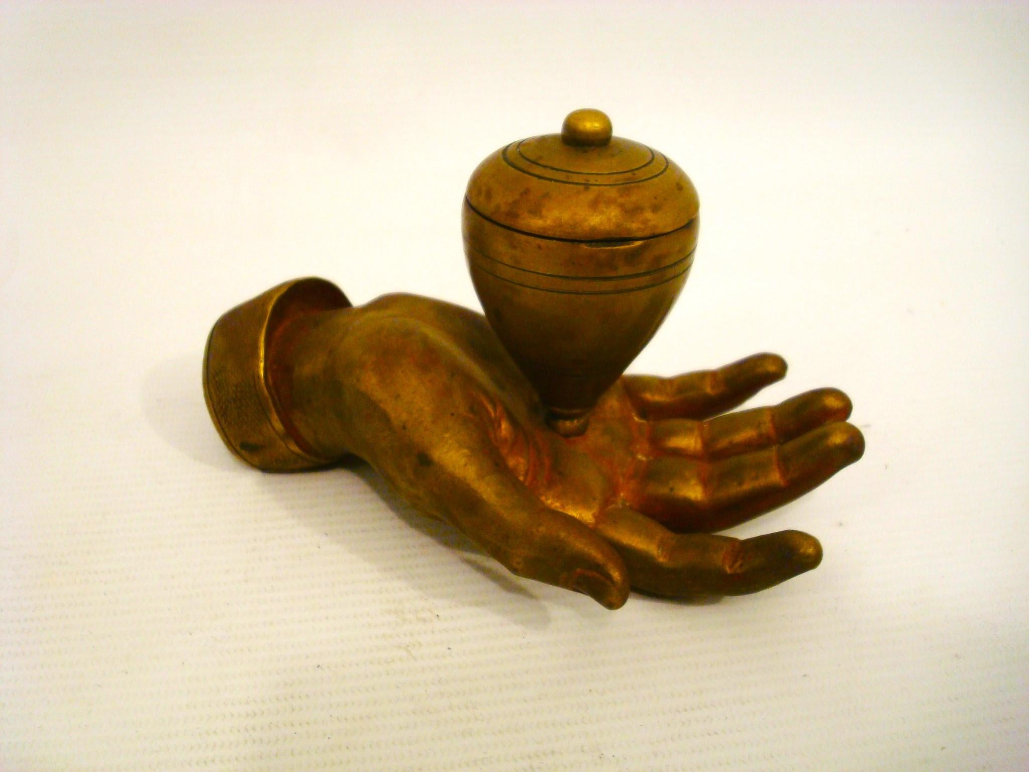 A bronze sculpture of a hand with an open palm. Resting on the palm is a  bronze object that resembles a traditional spinning top. The hand appears to be life-sized (Small Children size) and detailed, with visible lines and texture that mimic a real