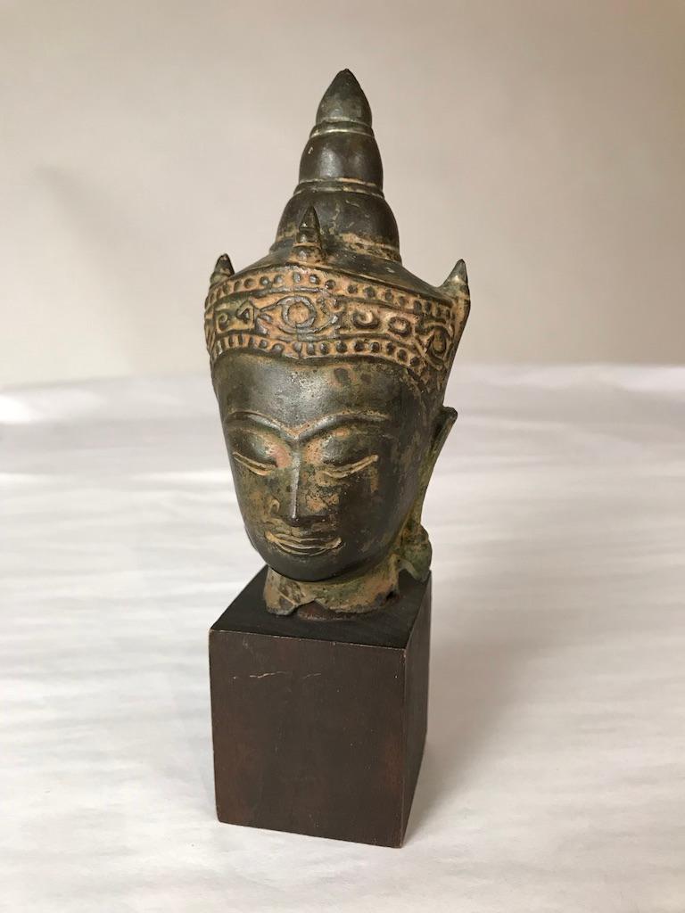 A Thai bronze head of Buddha, Sukothai period, late 16th-early 17th century. His face with serene expression, arched eyebrows above incised eyes and lips, aquiline nose. Wearing a decorated tiara in front of a conical-shaped usnisha. Mounted on a
