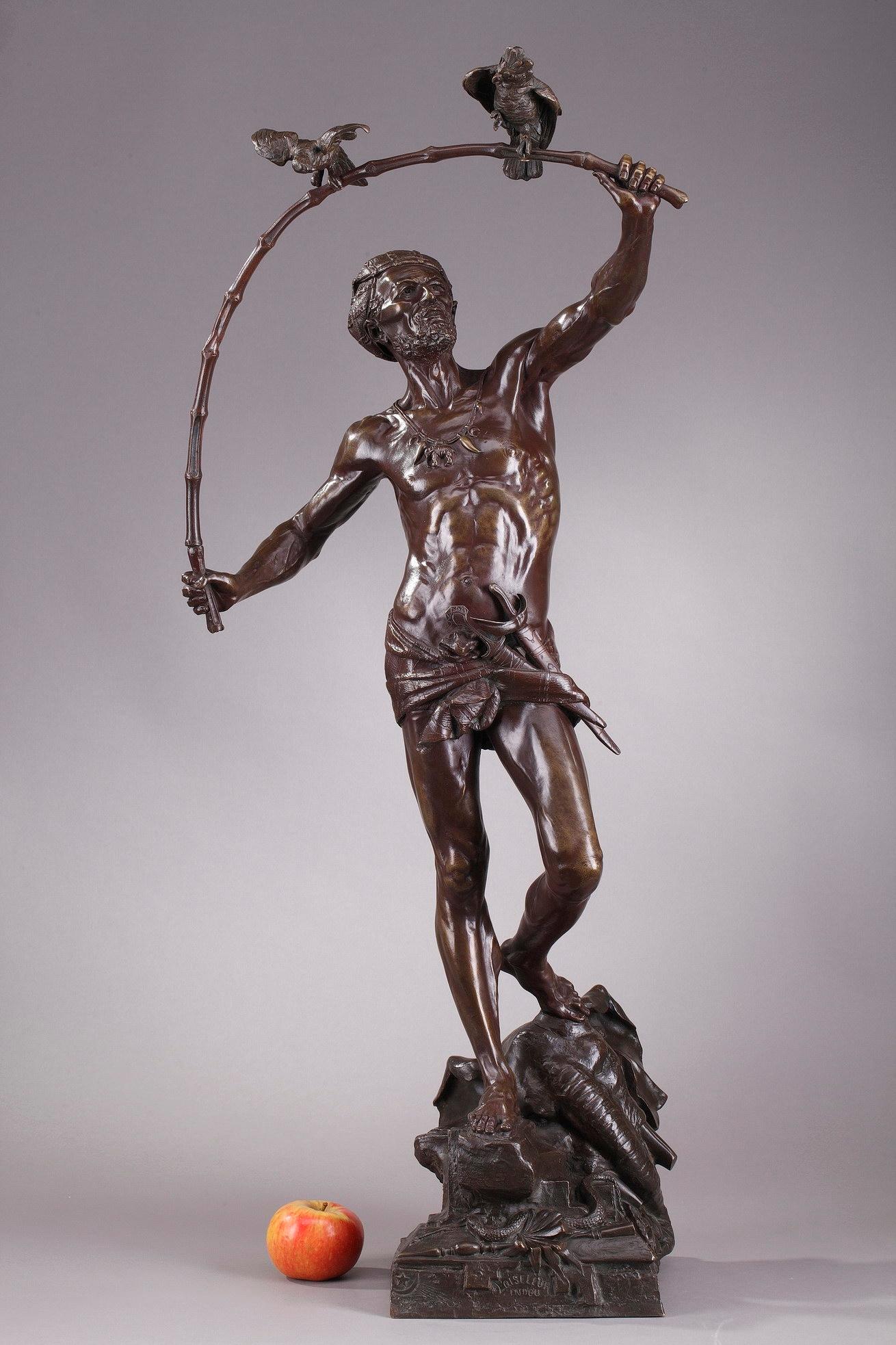 Grande statua in bronzo raffigurante un cacciatore di uccelli indù, opera dello scultore e medaglista belga Auguste de Wever. An He indossa un nastro e un drappeggio che sostiene una piccola spada. La sua collana è ornata da un medaglione a forma di