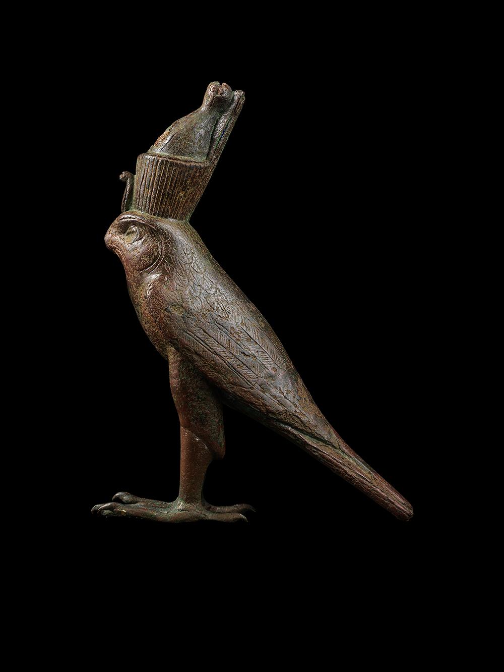 A bronze Horus in the form of a falcon, wearing the double crown of Upper and Lower Egypt, with a frontal uraeus. The wings are folded down the back of the falcon. The feet end with long talons, and sit on two vertical projections, which would have