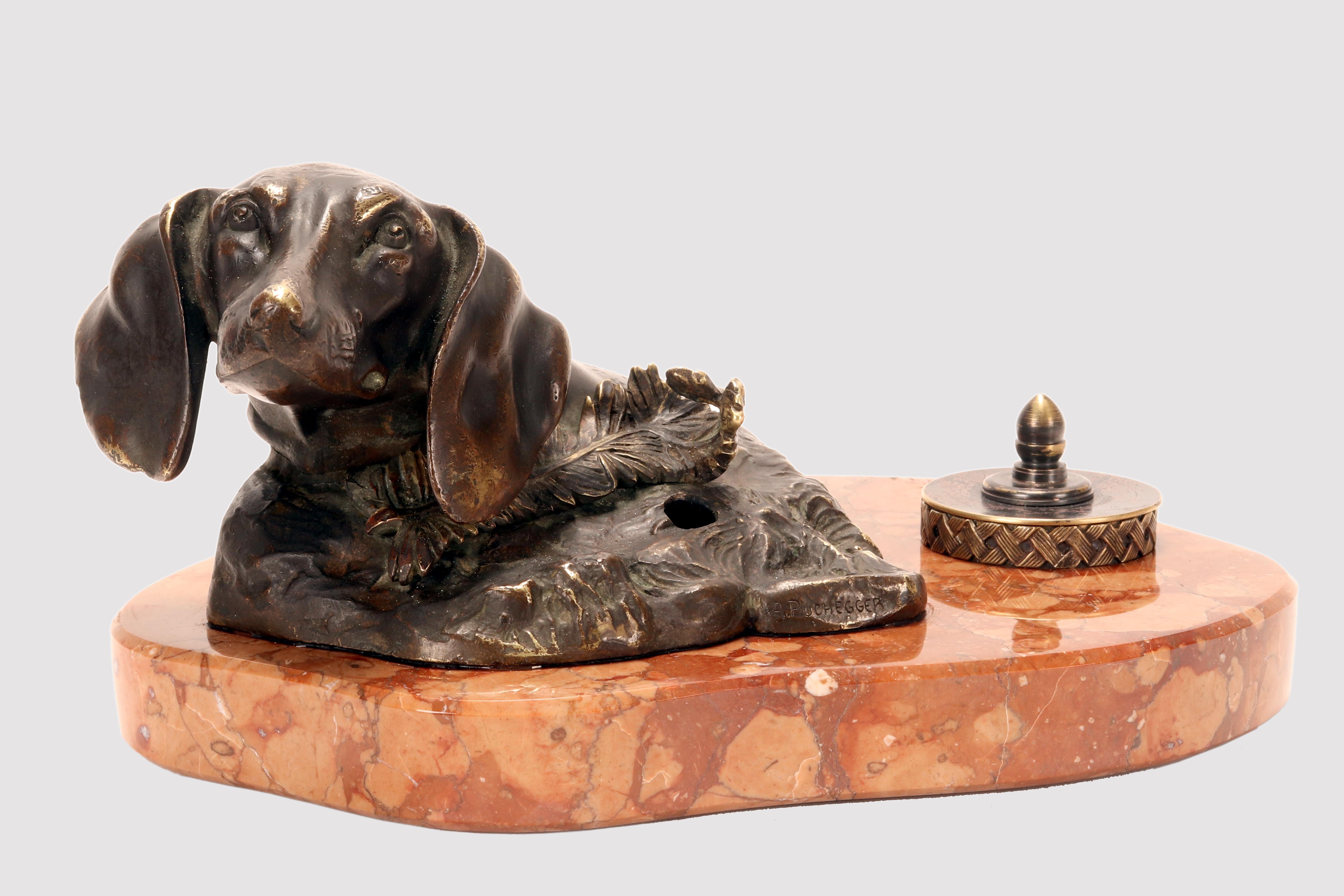 Bronzetisch-Tintenfass mit Federhalter, der den Kopf eines Dackels darstellt. Die Skulptur ruht auf einem geschwungenen Sockel aus rotem Veroneser Marmor, der auch den Tintenbehälter aus Messing aufnimmt. Unterzeichnet A. Puchegger (1878-1917).