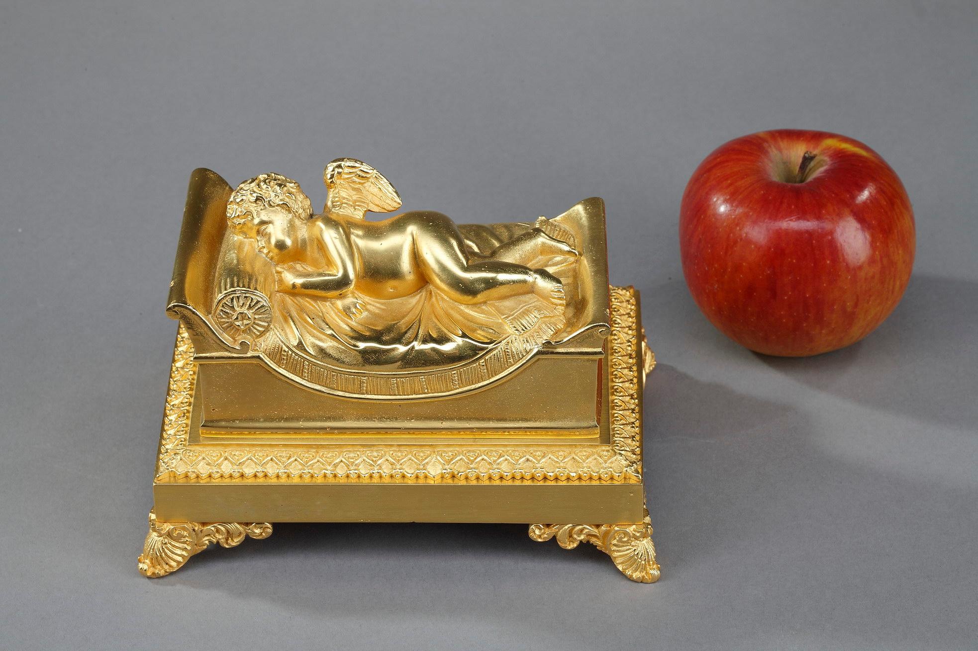 Small bronze inkwell dating from the first part of the 19th century. The lid of the inkwell represents a bed on which a putto is dozing. The whole rests on a rectangular base decorated with a frieze of ovals with four feet in leafy hooves. It is