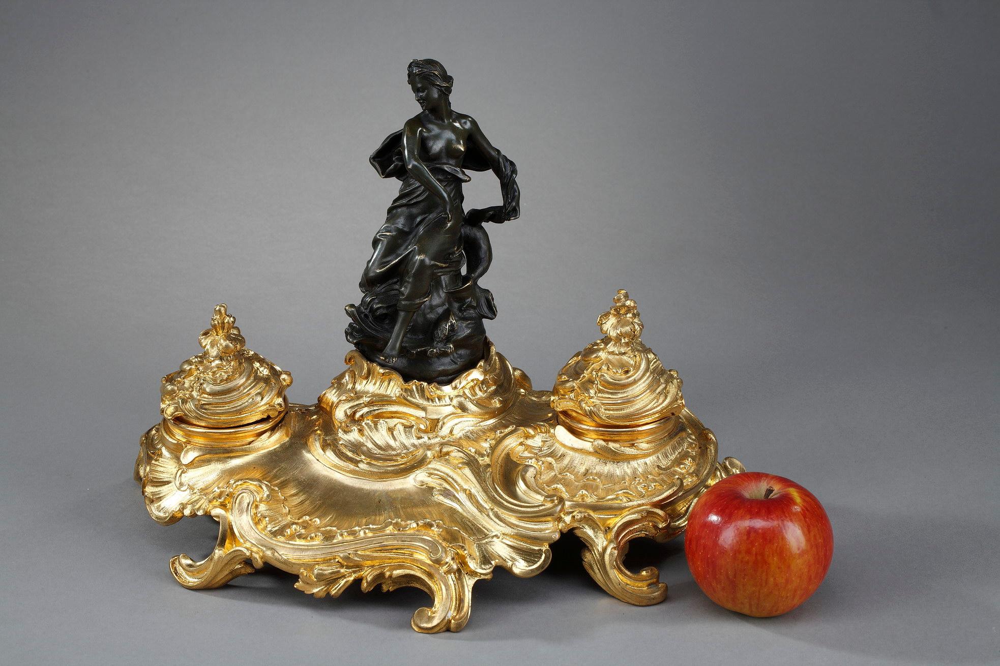 Rocaille inkwell representing a goddess in the Antique style in bronze with brown patina, resting on a gilt bronze base decorated with rocaille and foliage scrolls, with two cups. The lids of the inkwells are chiseled with leaves. The deep work