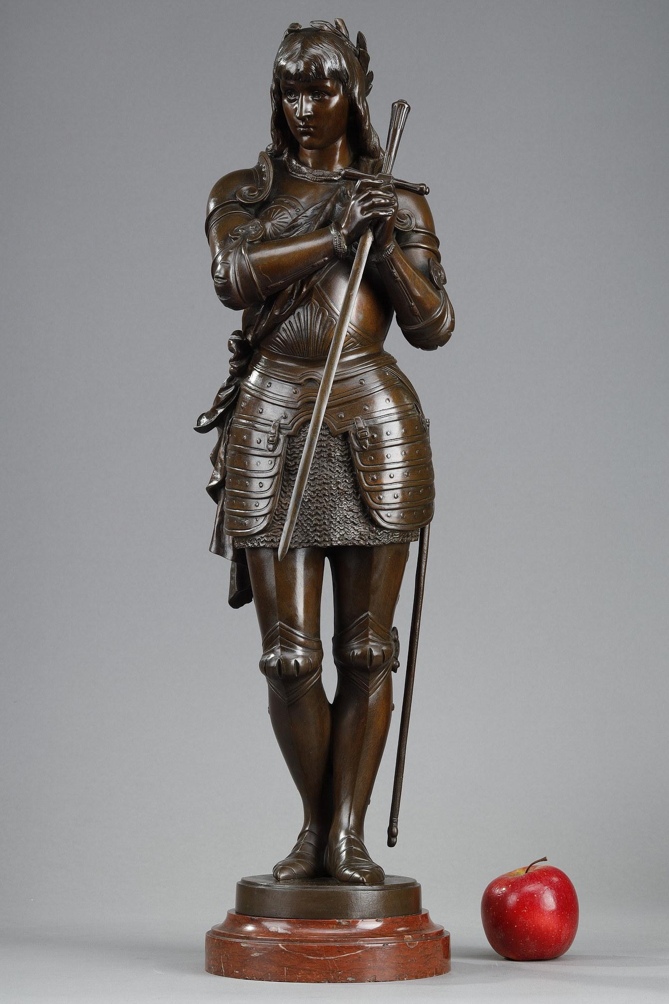 Brown patina bronze sculpture representing Joan of Arc signed Eutrope Bouret. The saint is depicted standing, dressed in medieval armour with a scarf tied around her waist, her hands joined in prayer around her sword. This gesture parallels her role