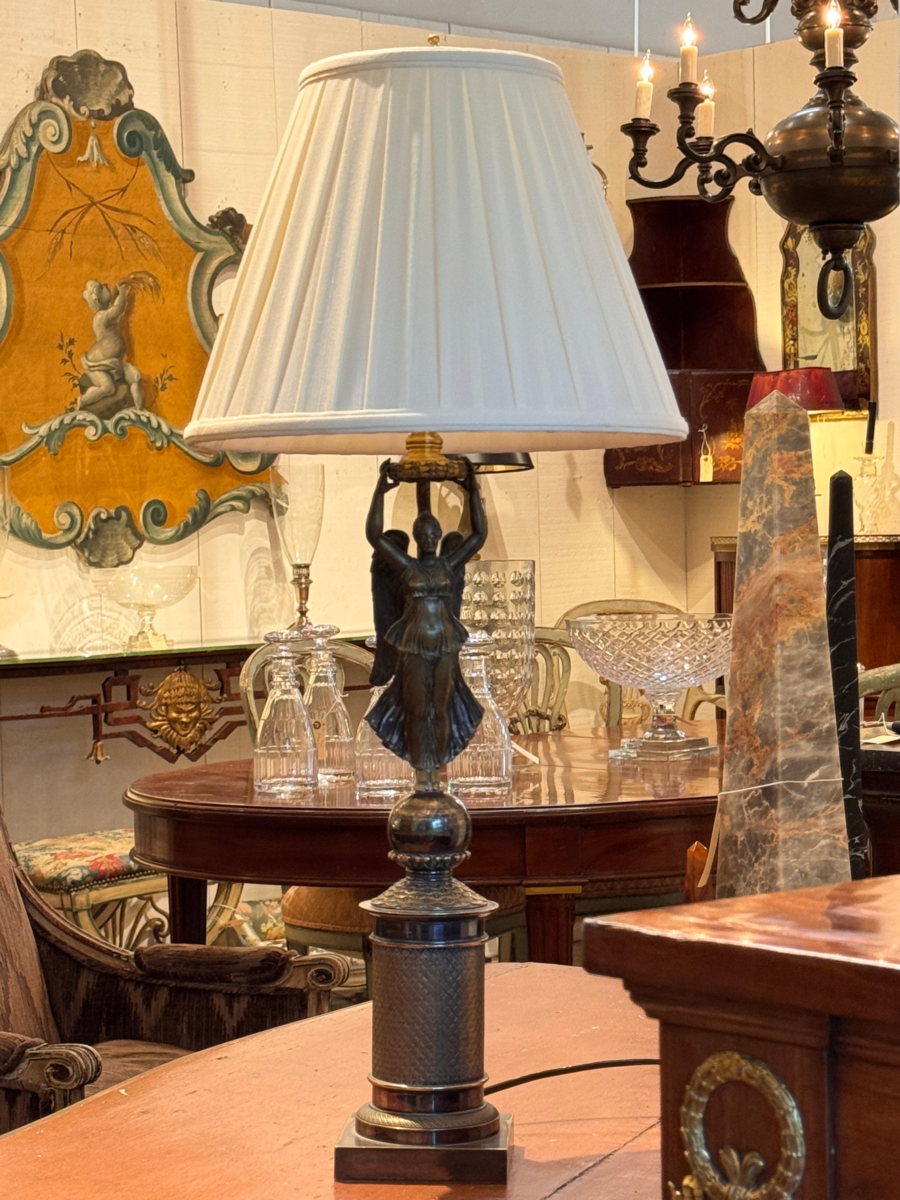 Elegant bronze lamp featuring a finely cast winged female figure atop a cylindrical base with textured detailing and stepped plinth. The classical form and patinated finish highlight the lamp’s neoclassical inspiration. The close-up view reveals