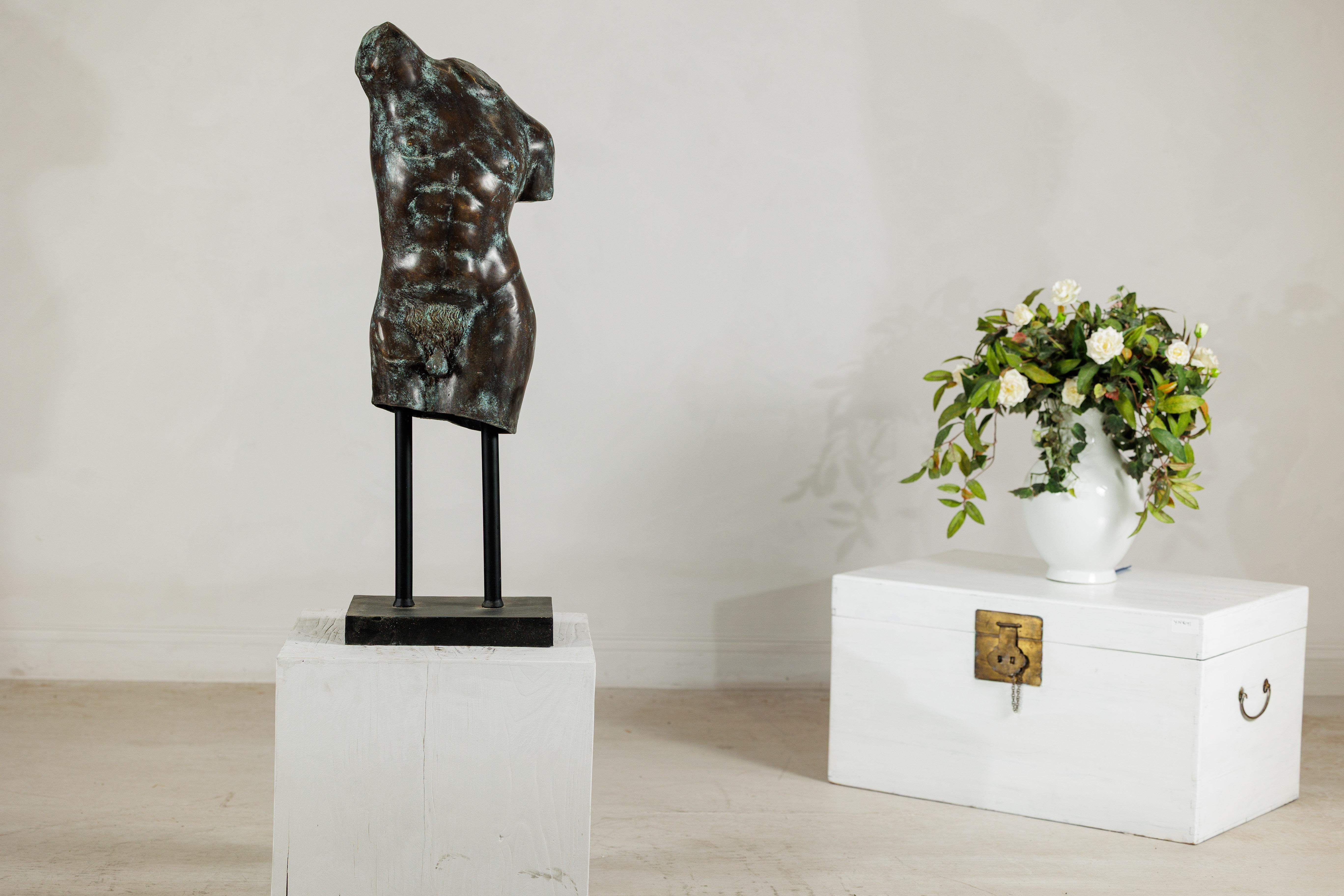 A bronze male torso sculpture presented on a custom stand, its fragmented classical form evoking the timeless beauty of antiquity. The figure, modeled with well-defined musculature and naturalistic detail, is rendered without head or limbs,