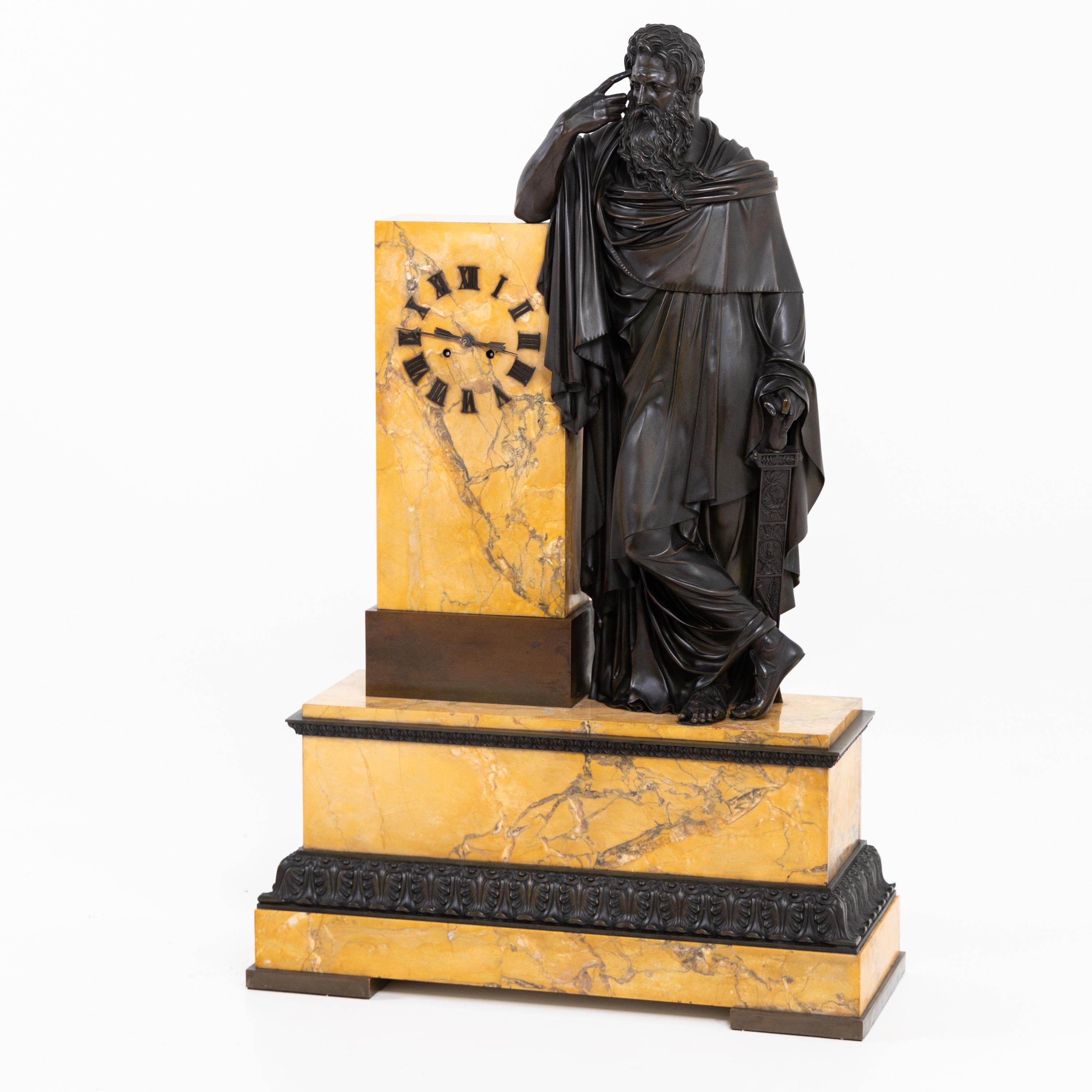 La grande pendule sur un socle en marbre de Sienne avec frise en bronze patiné ainsi que la grande représentation entièrement sculptée d'un homme barbu avec l'index à la tempe. L'autre main repose librement sur le pommeau d'une épée. Le mouvement